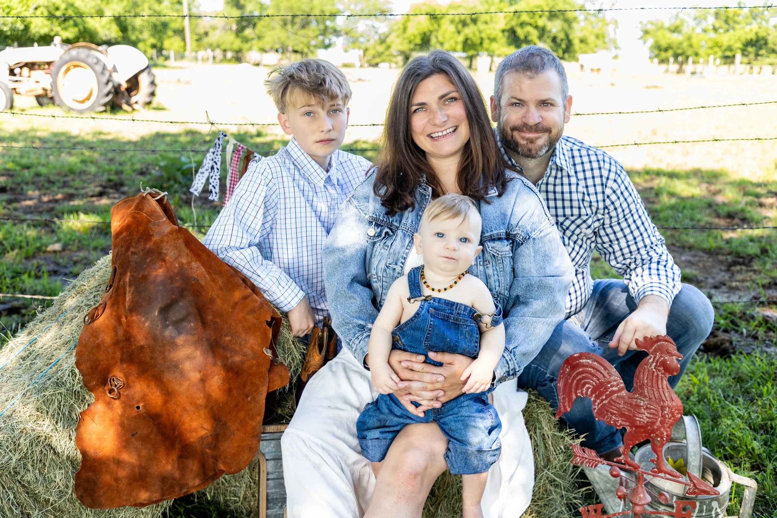 A family of four posing outdoors in a rural setting with a tractor in the background, sitting on hay bales surrounded by farm decorations.