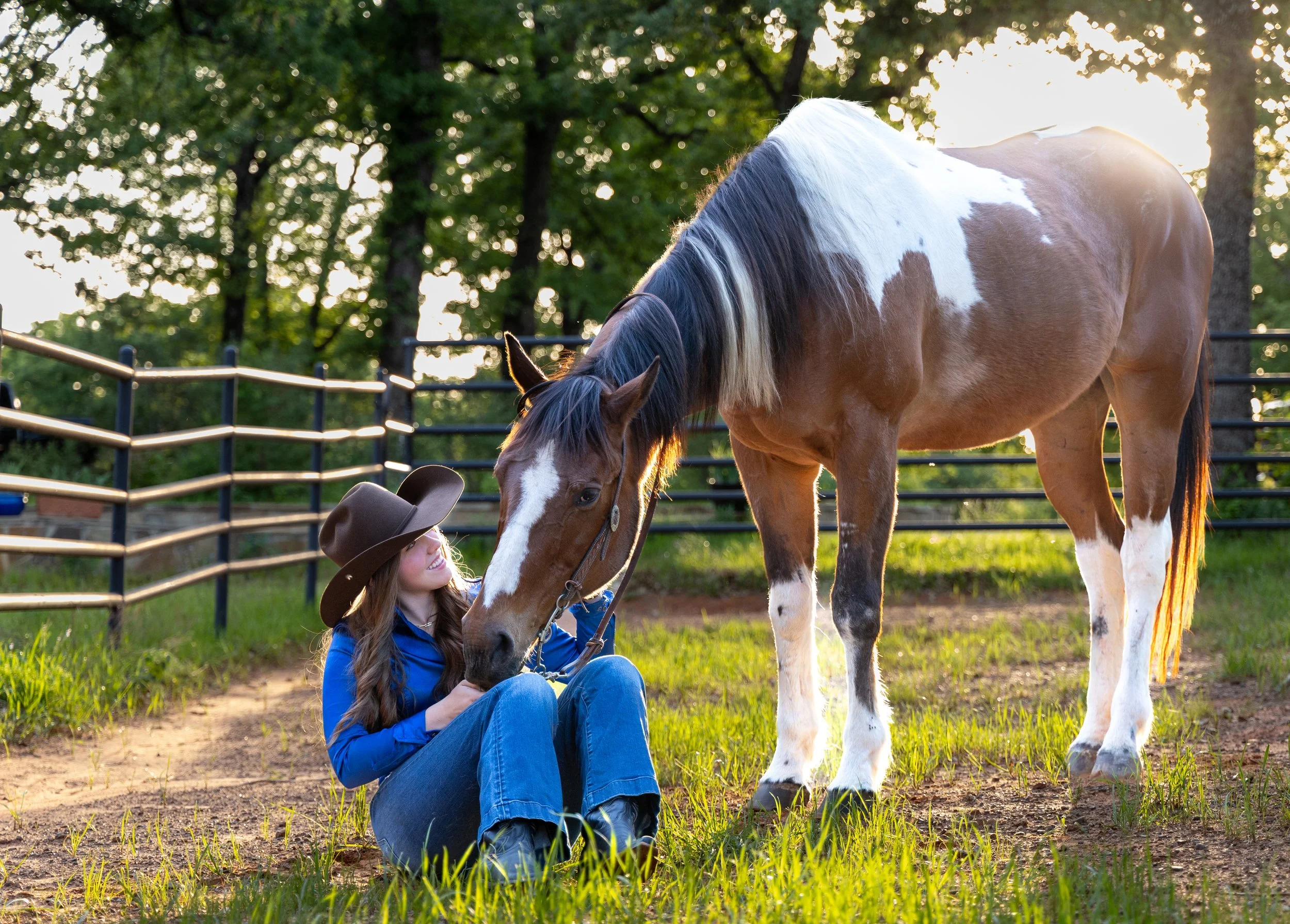 A girl sitting on the grass, smiling, with a brown and white horse. The girl is wearing a blue jacket and a cowboy hat, and the horse has a bridle.