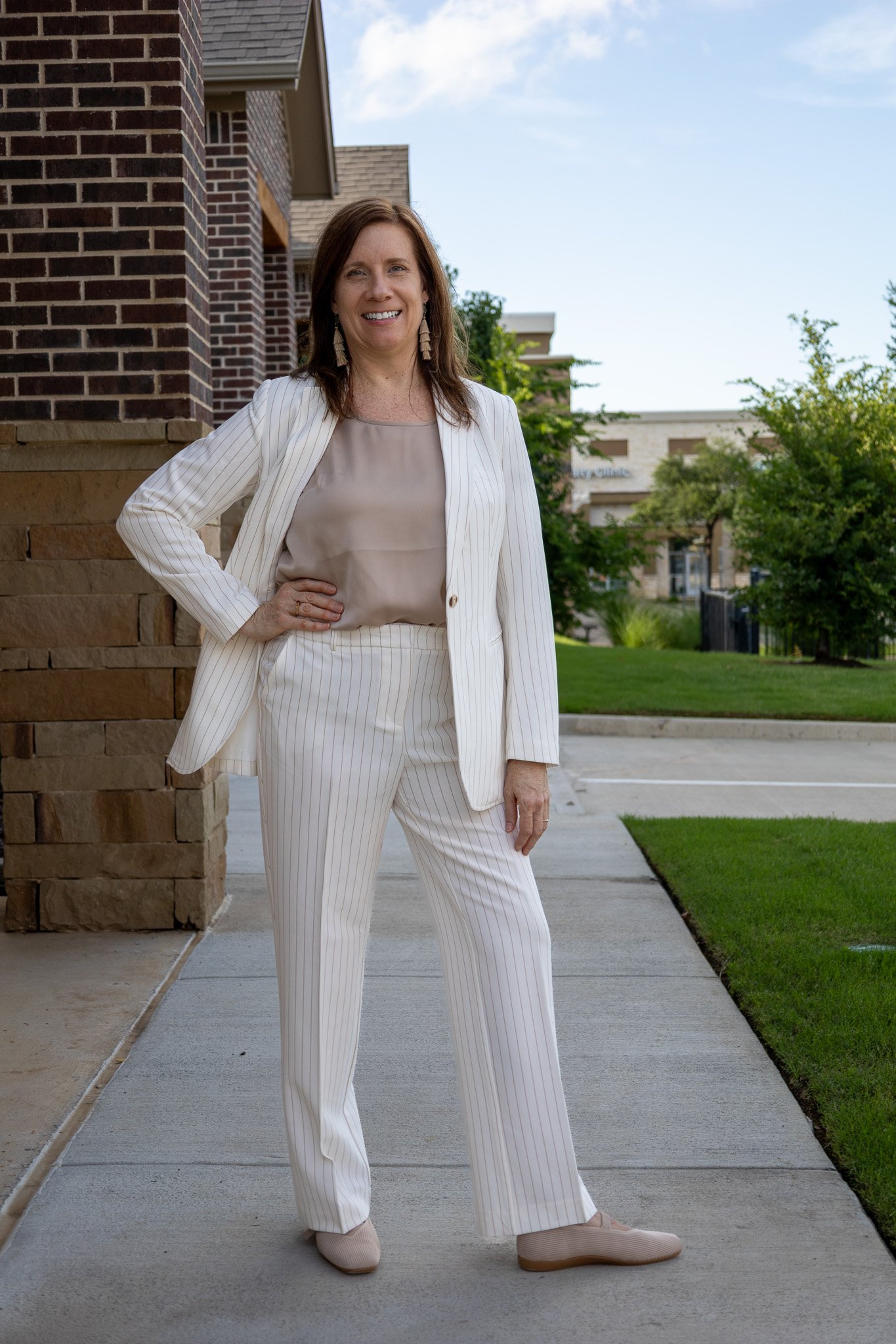 Woman standing outside on sidewalk, smiling, wearing a white striped blazer and matching pants, beige blouse, and beige shoes.