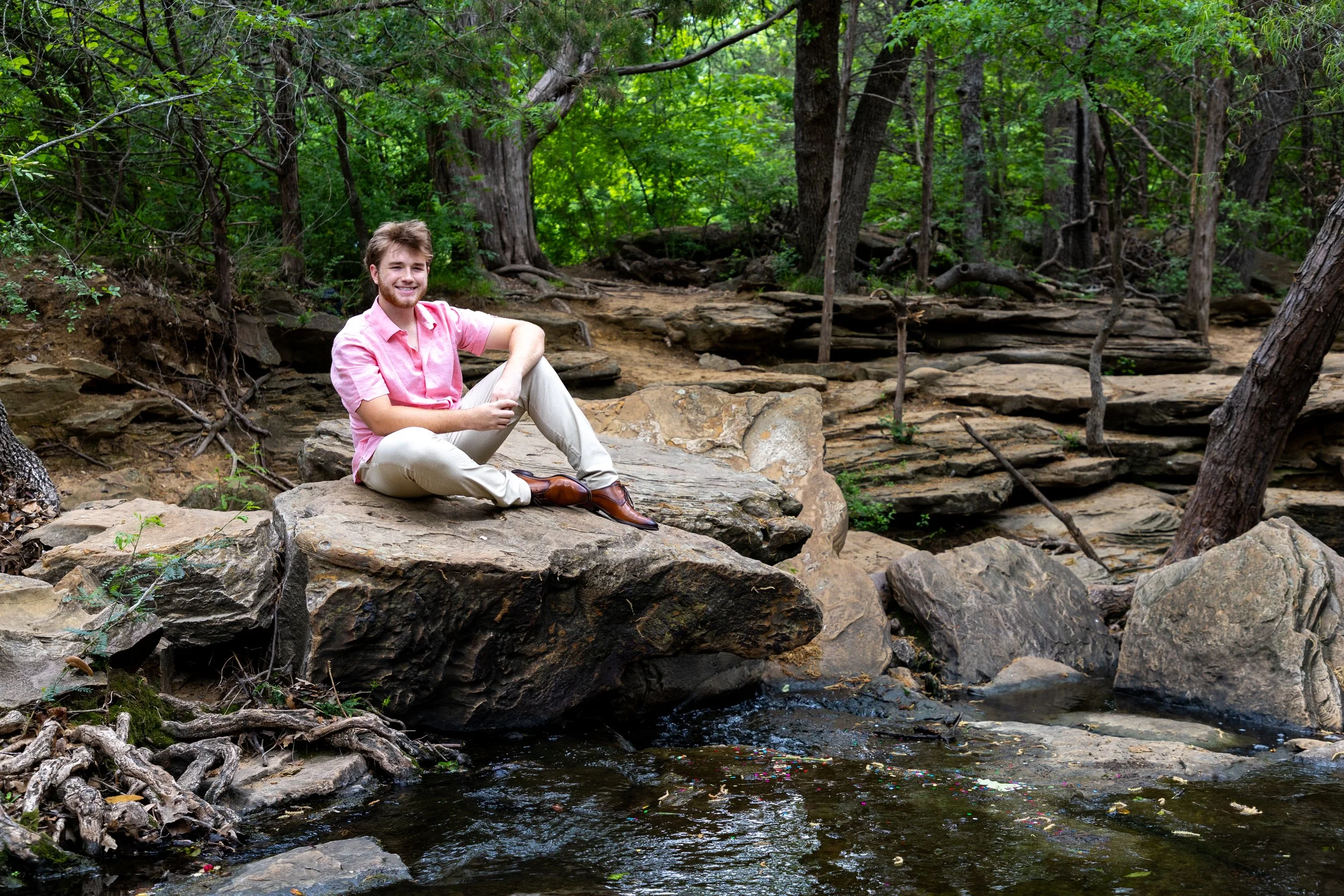 A man sitting on a large rock by a creek in a forest, smiling at the camera, surrounded by trees and greenery.