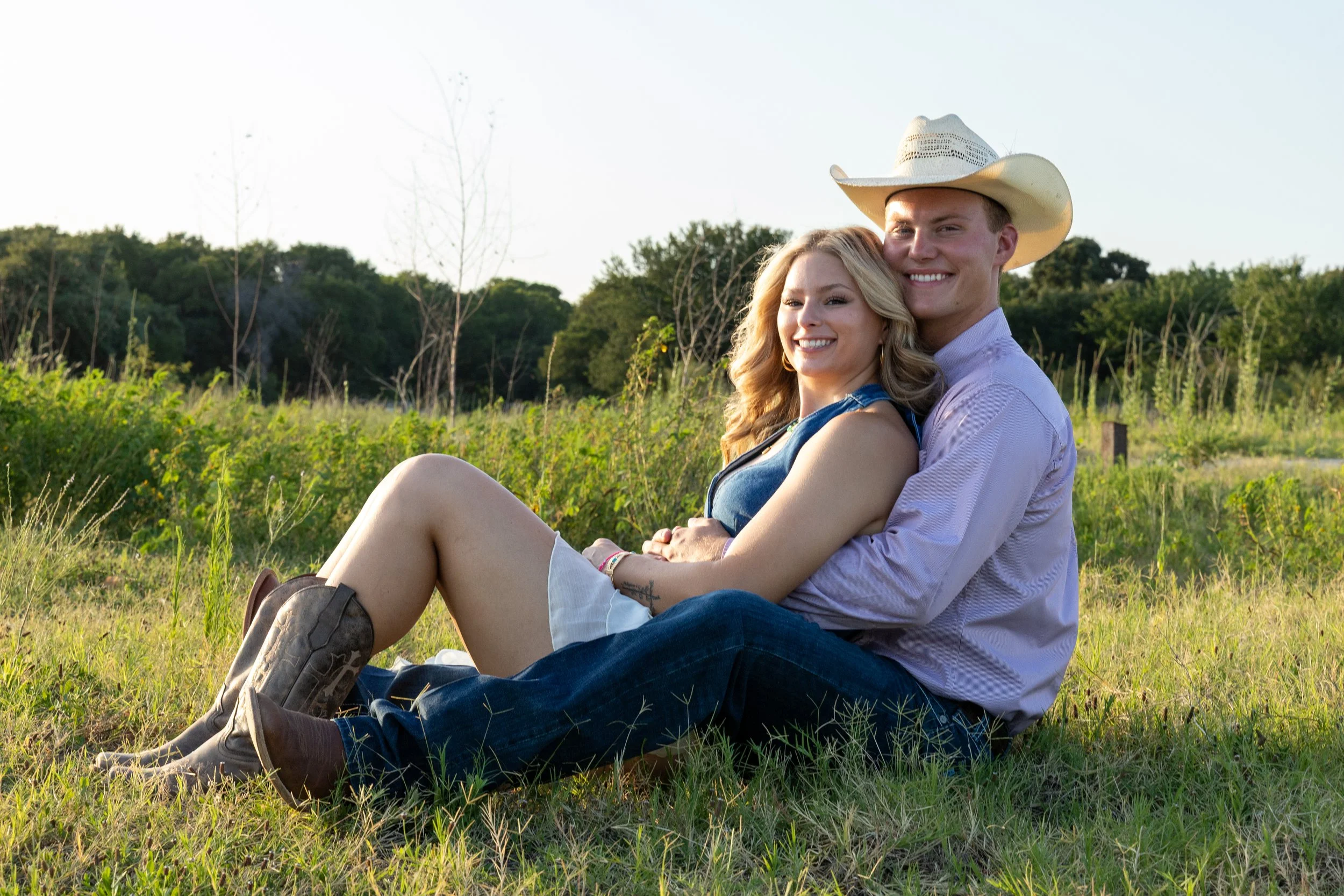 A young couple sitting on grass in a field, smiling at the camera, the man wearing a cowboy hat and the woman leaning against him.