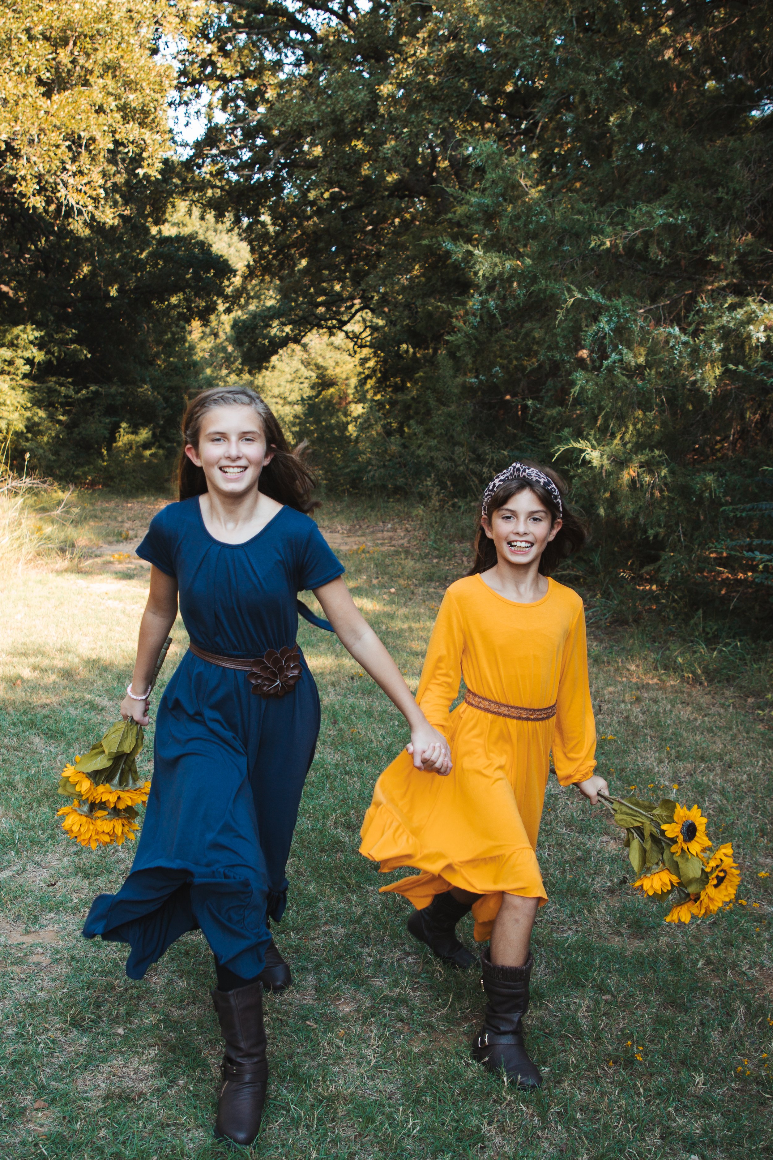Two young girls in colorful dresses holding sunflowers and walking outdoors in a grassy area with trees in the background.