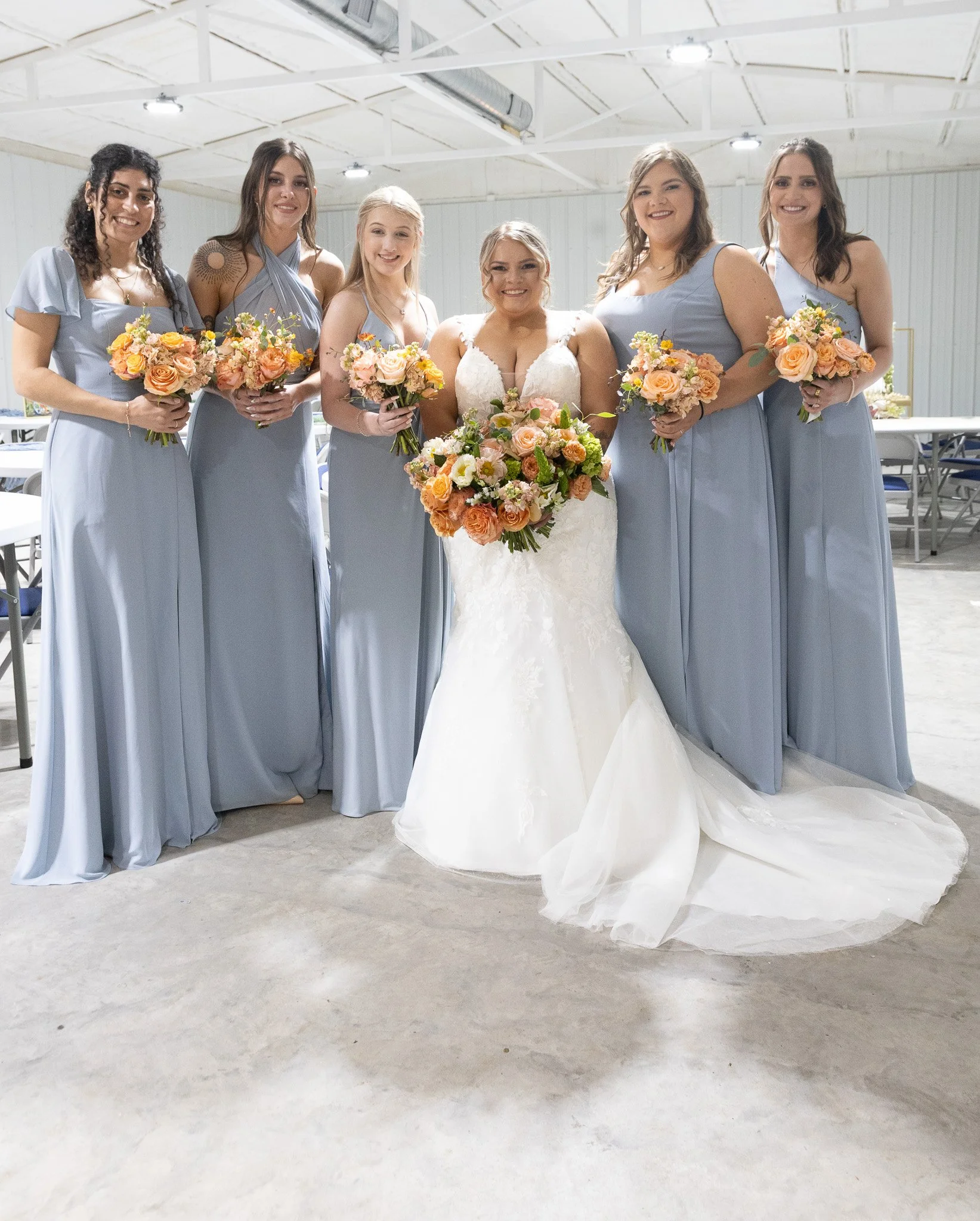 Bride in a wedding dress standing with six bridesmaids in light blue dresses, all holding bouquets of flowers, inside a large industrial-style venue.
