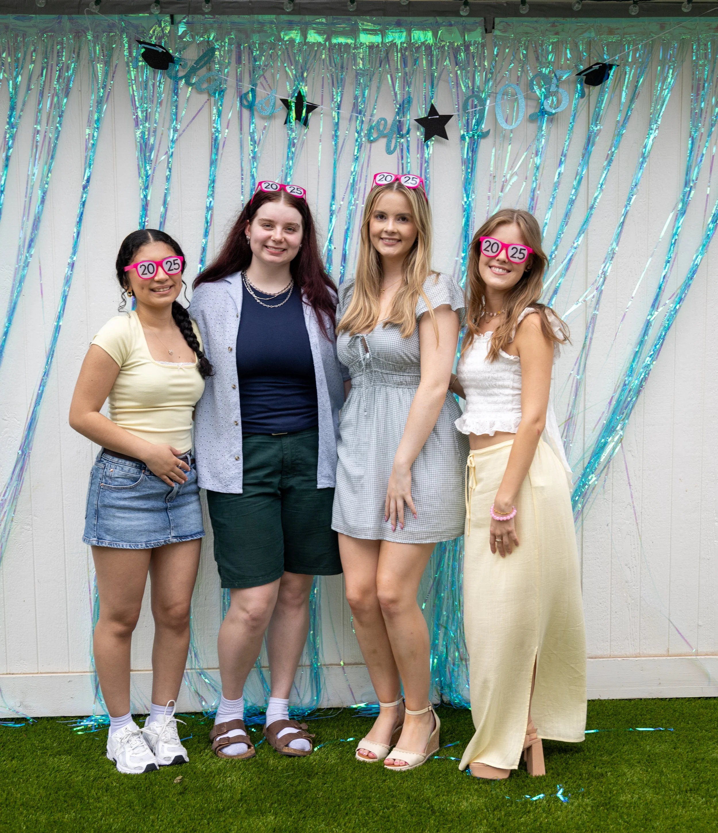 Four young women at a graduation celebration, wearing glasses with '20' and '25' labels, standing in front of a shiny, iridescent backdrop with graduation caps and stars decorations.