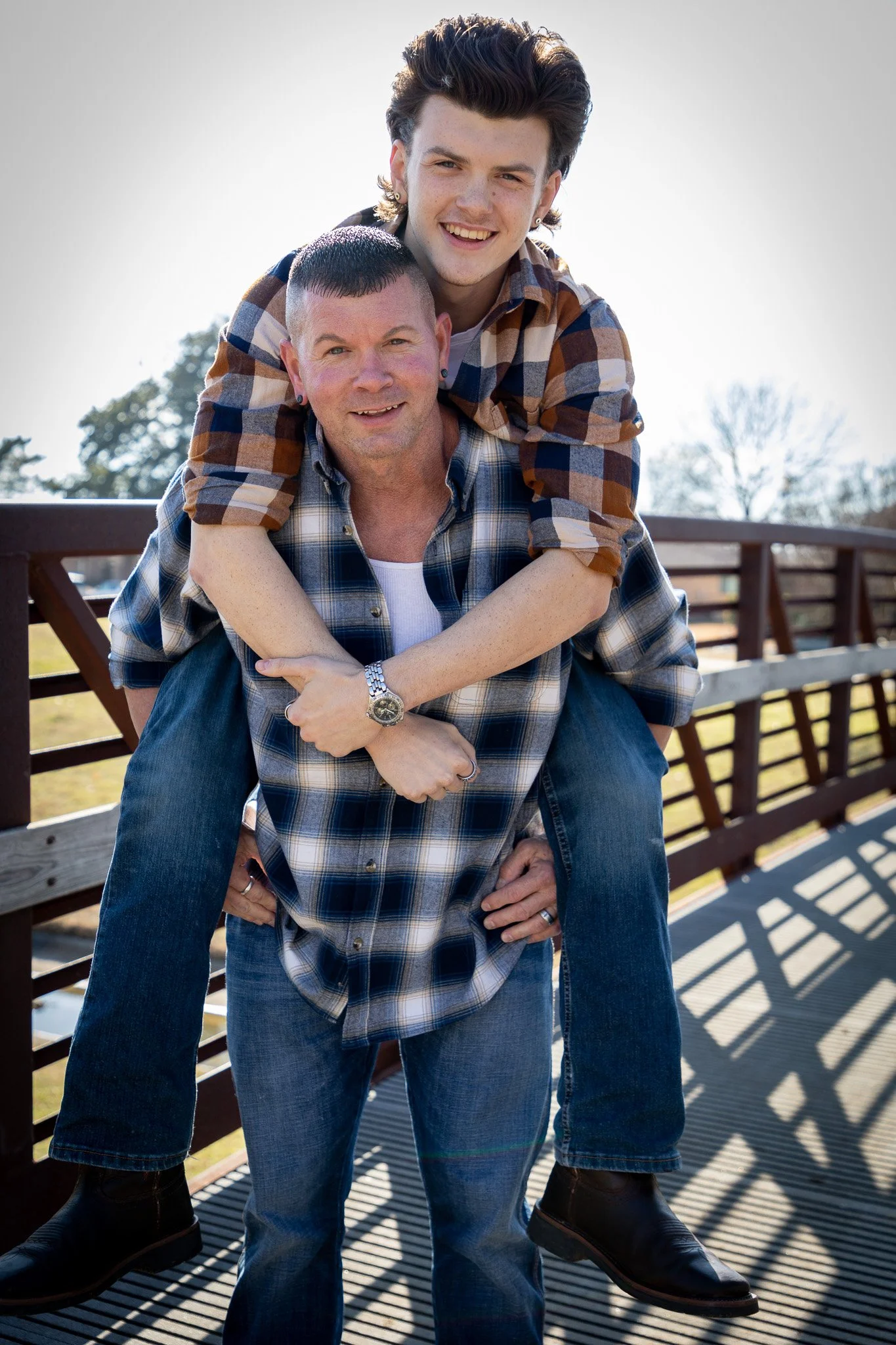 Father and son outdoors on a wooden bridge, the son sitting on the father's shoulders with arms around his father's neck, smiling at the camera.