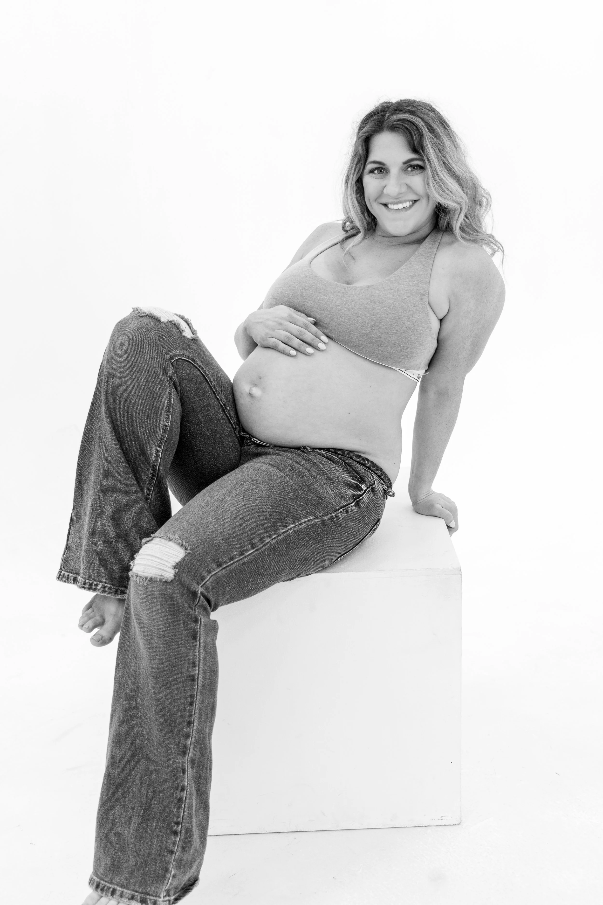 Black and white photo of a pregnant woman sitting on a block, smiling, wearing a tank top and ripped jeans.