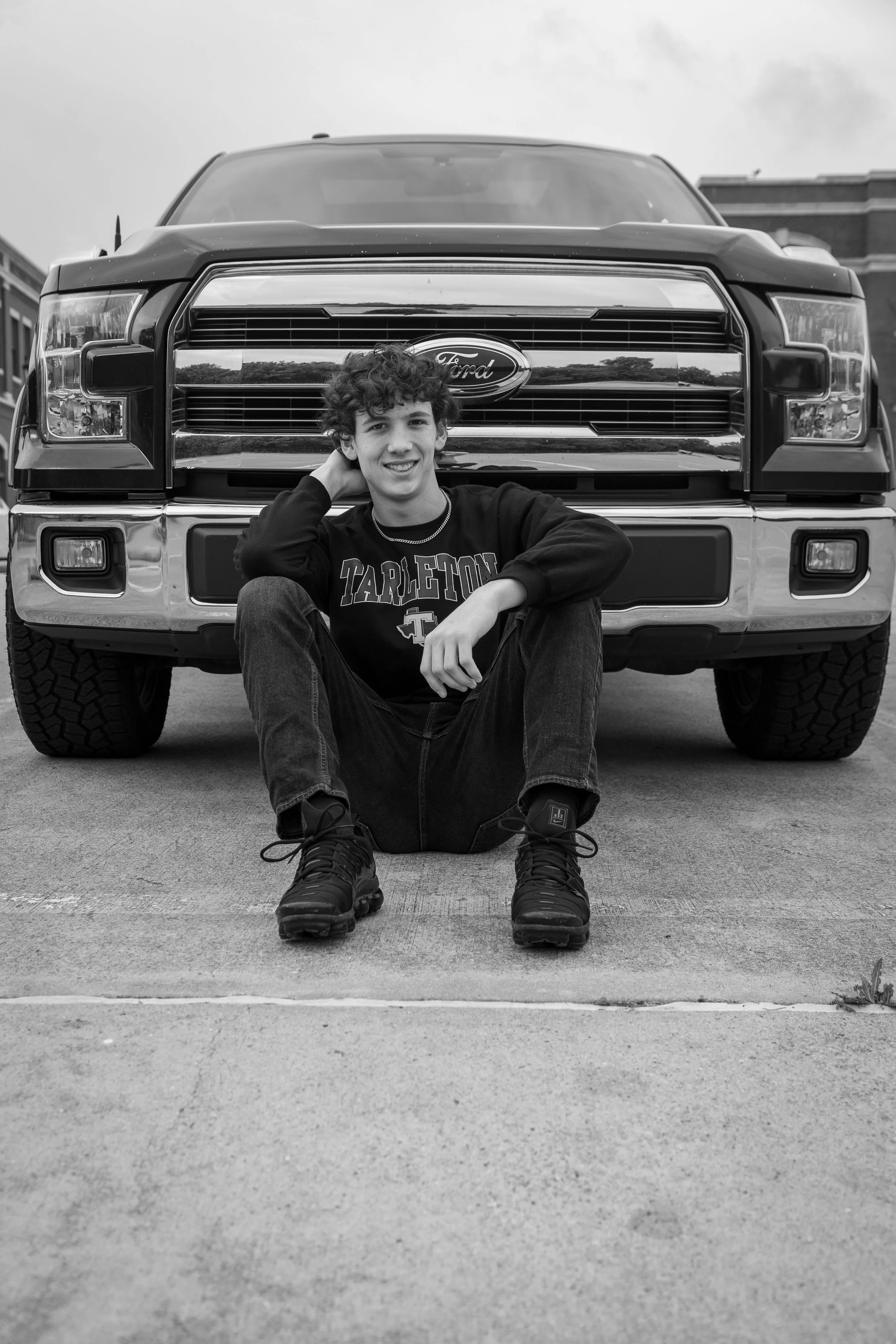 A young man sitting on the ground in front of a large Ford truck, smiling at the camera, with his hand resting behind his head.