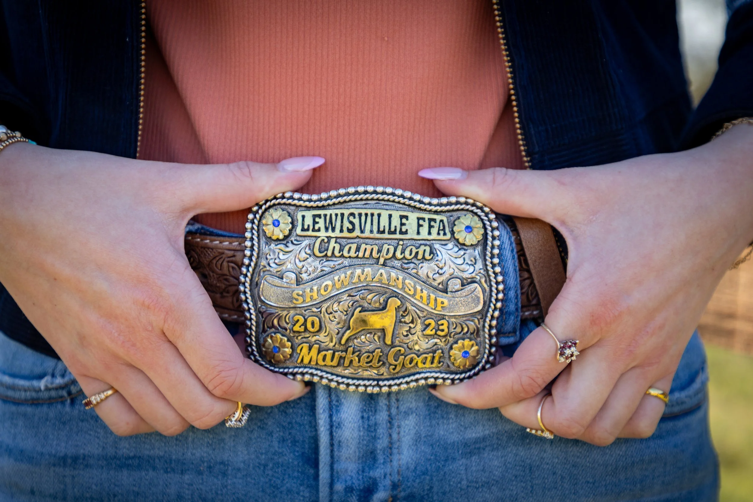 Close-up of a person holding a championship belt with the text 'Louisville FFA Showmanship 2023 Market Goat' on it, wearing rings and a dark jacket with a pink top.