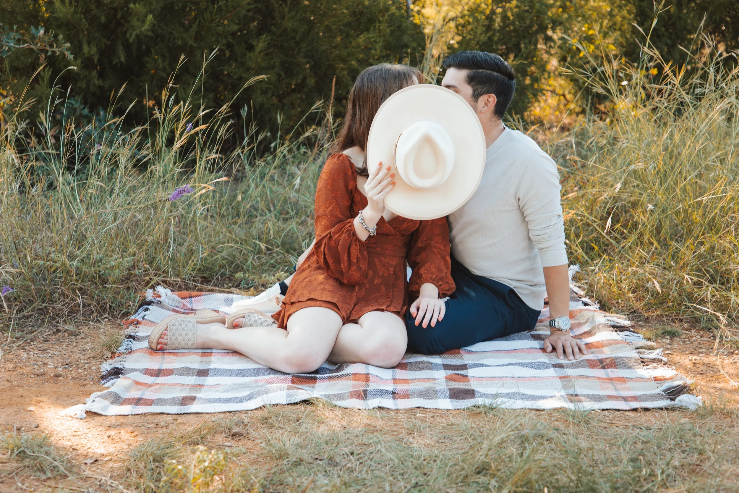 A couple sitting on a plaid blanket outdoors, with the woman holding a large white sunhat and both leaning in for a kiss hidden behind the hat amidst grassy surroundings and trees.