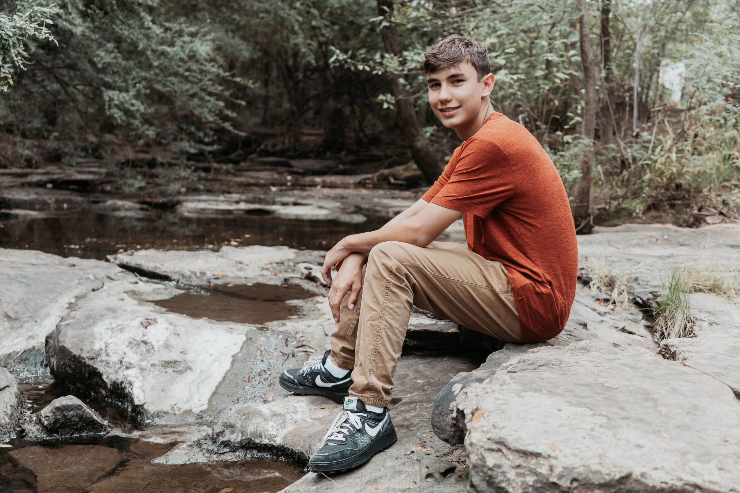 A teenage boy sitting on rocks by a small stream in a wooded area, smiling at the camera.