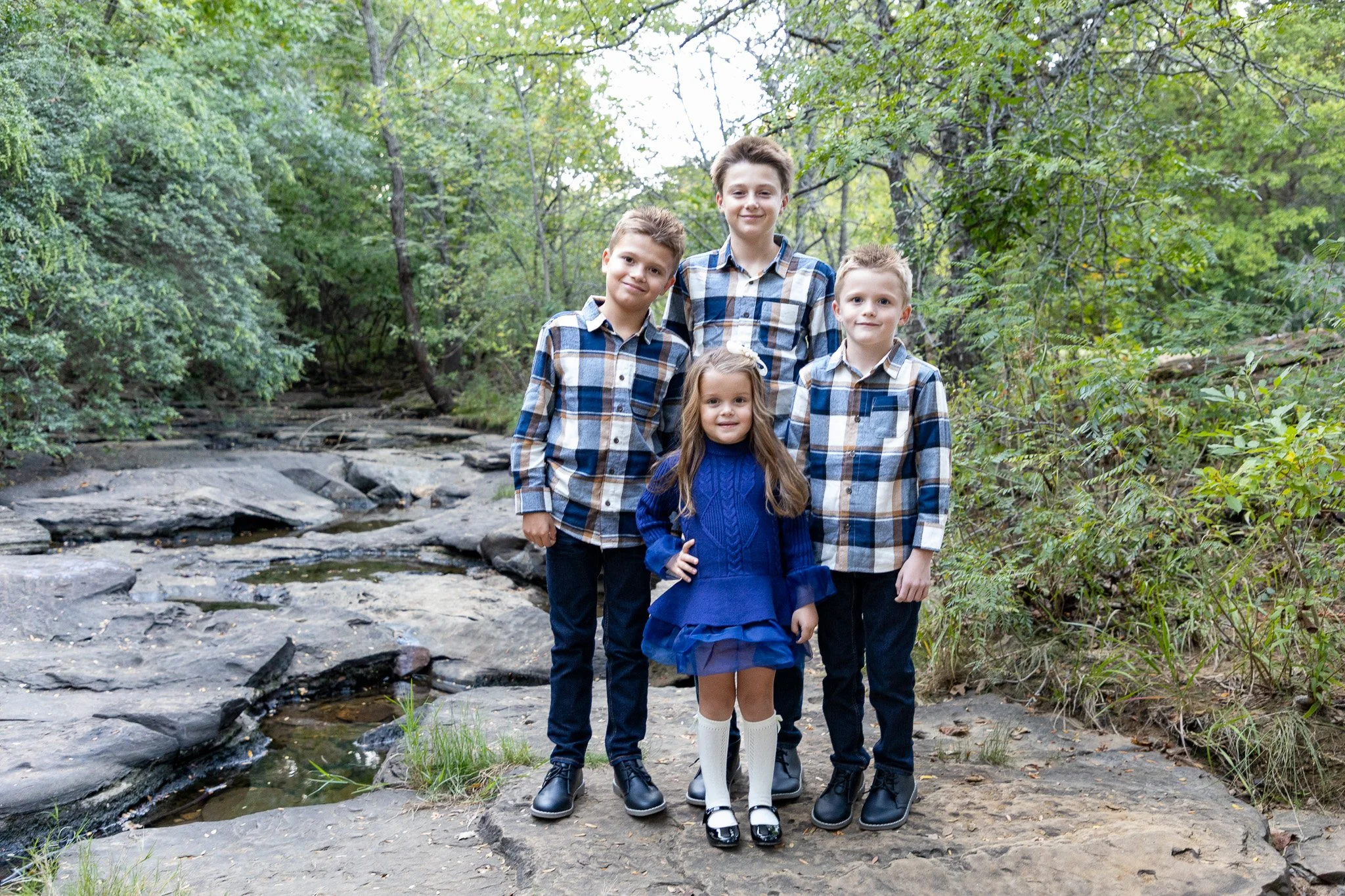 Four children, three boys and one girl, standing on rocks near a creek in a wooded area, dressed in casual clothing.