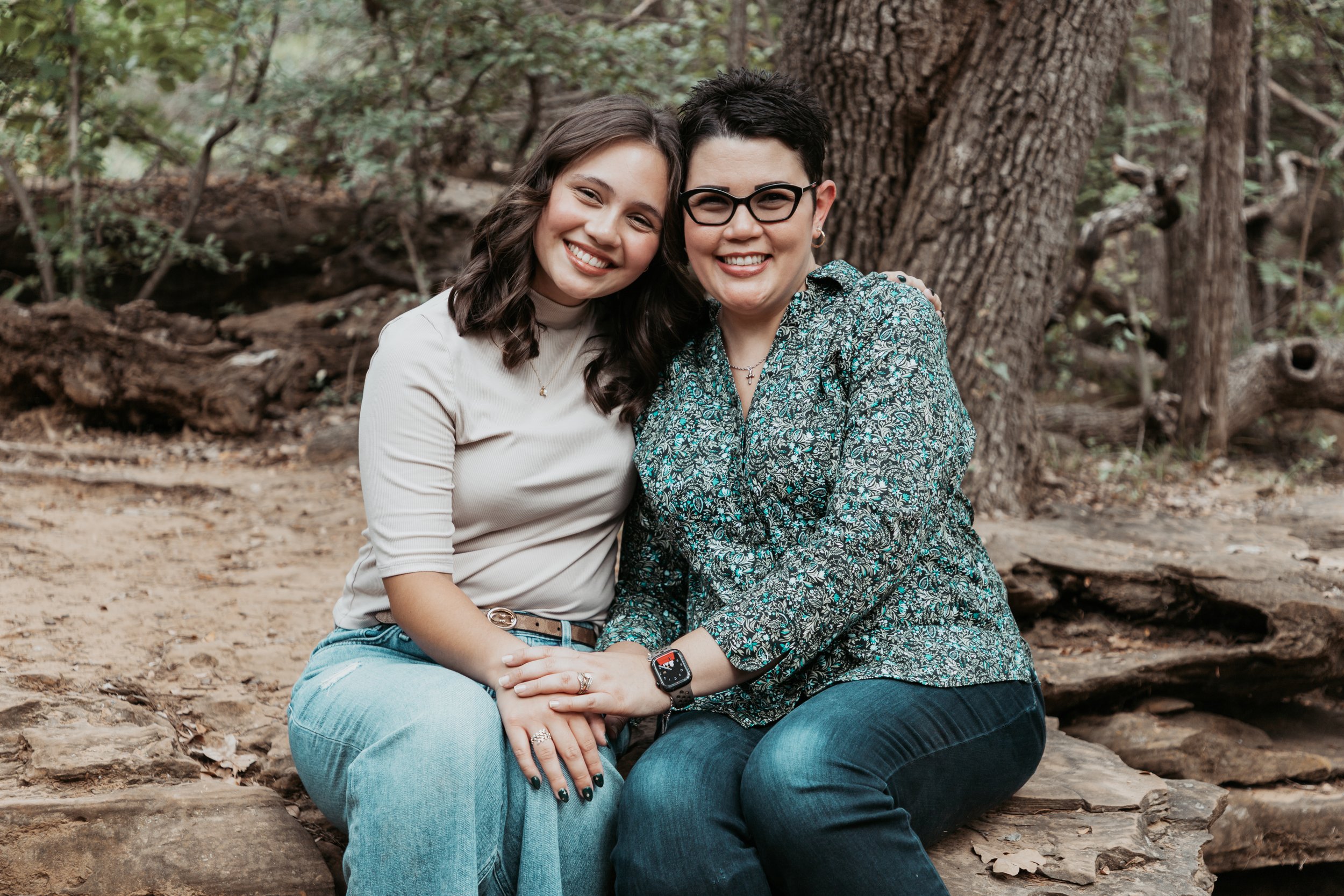 Two women sitting close together on a log in a wooded area, smiling at the camera.