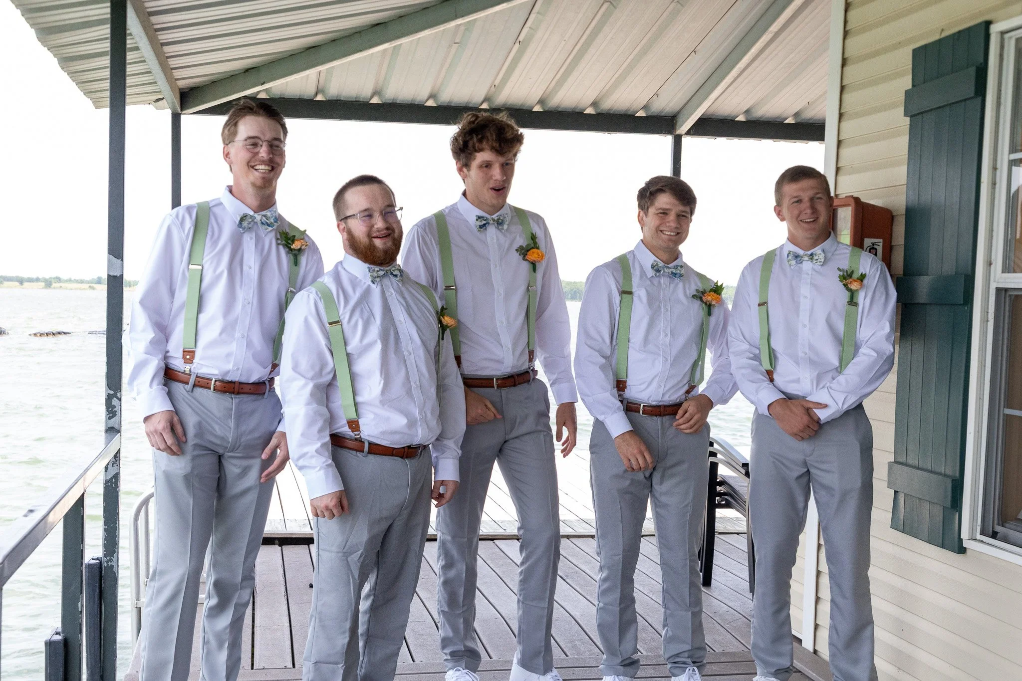 A group of six young men in dress shirts, suspenders, and bow ties standing on a porch overlooking a body of water, smiling and enjoying each other's company.