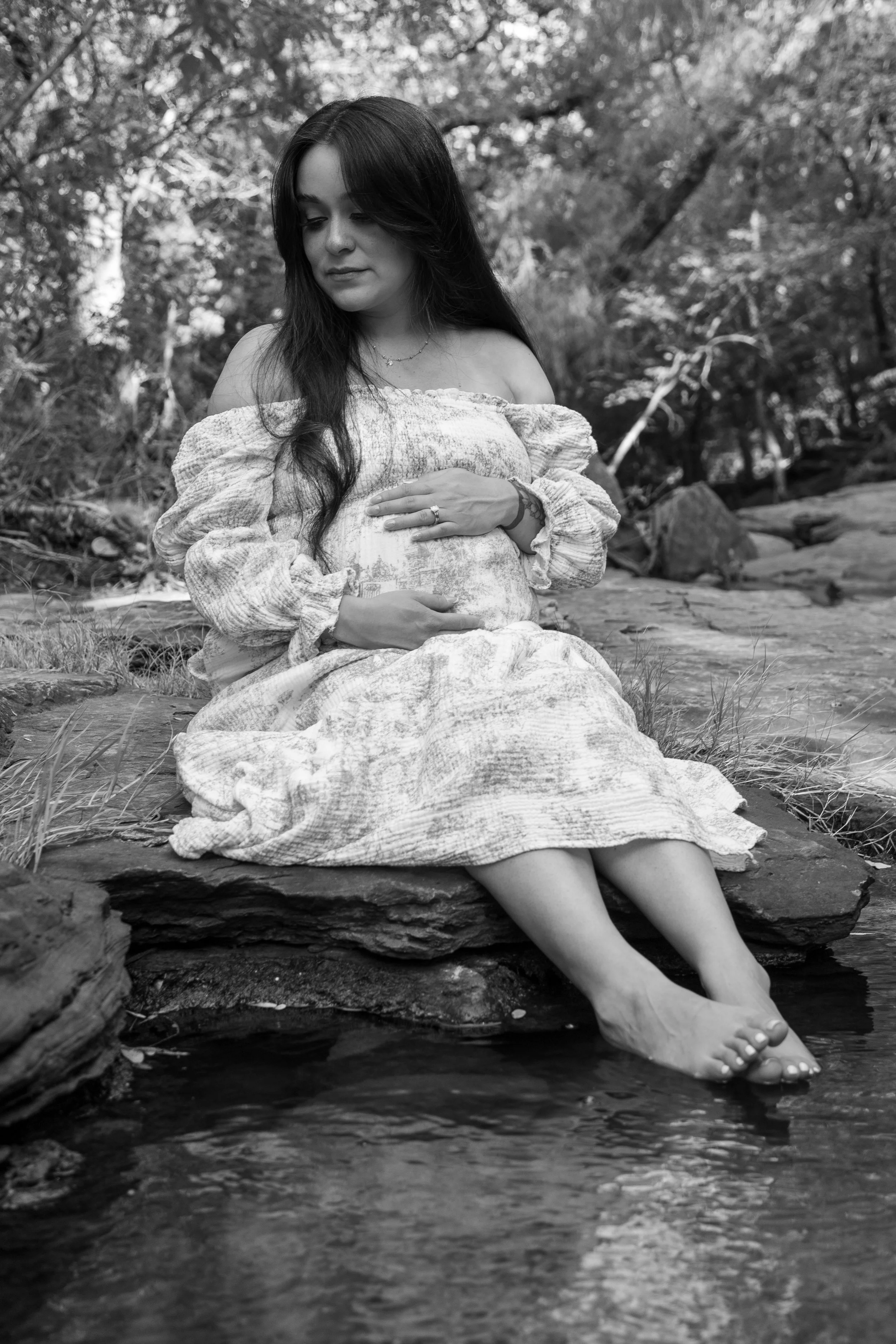 A woman sitting on a rock by a stream, with her feet in the water, surrounded by trees and nature, in black and white.