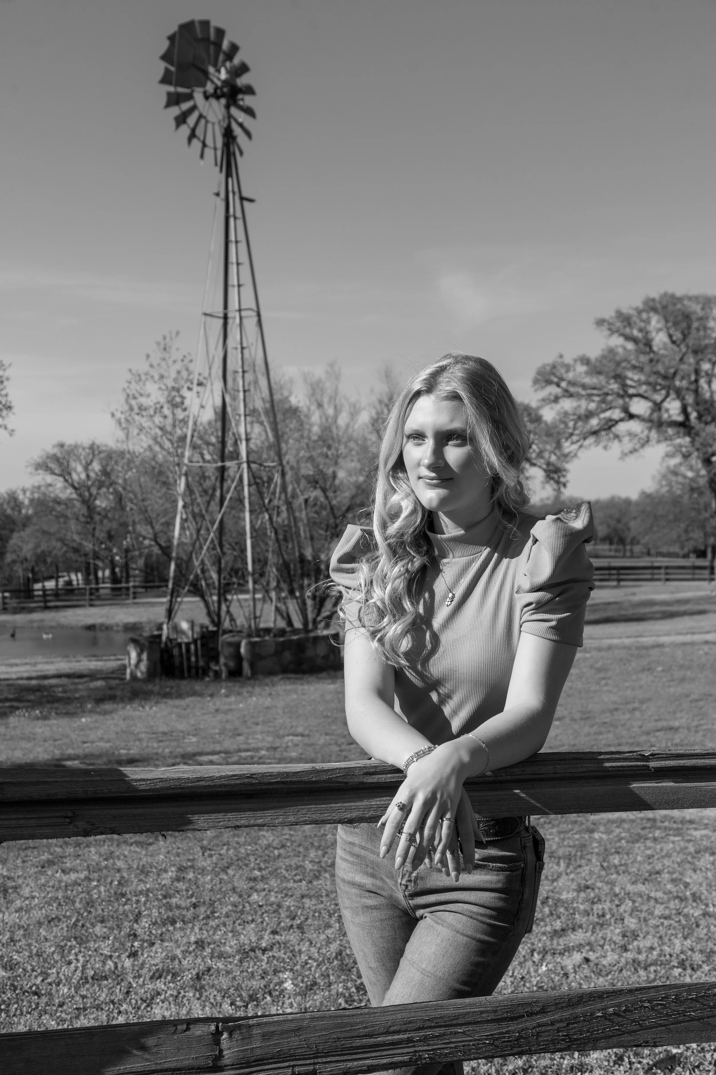 A young woman with long wavy hair leaning on a wooden fence in a park, with a windmill in the background, trees, and open sky.