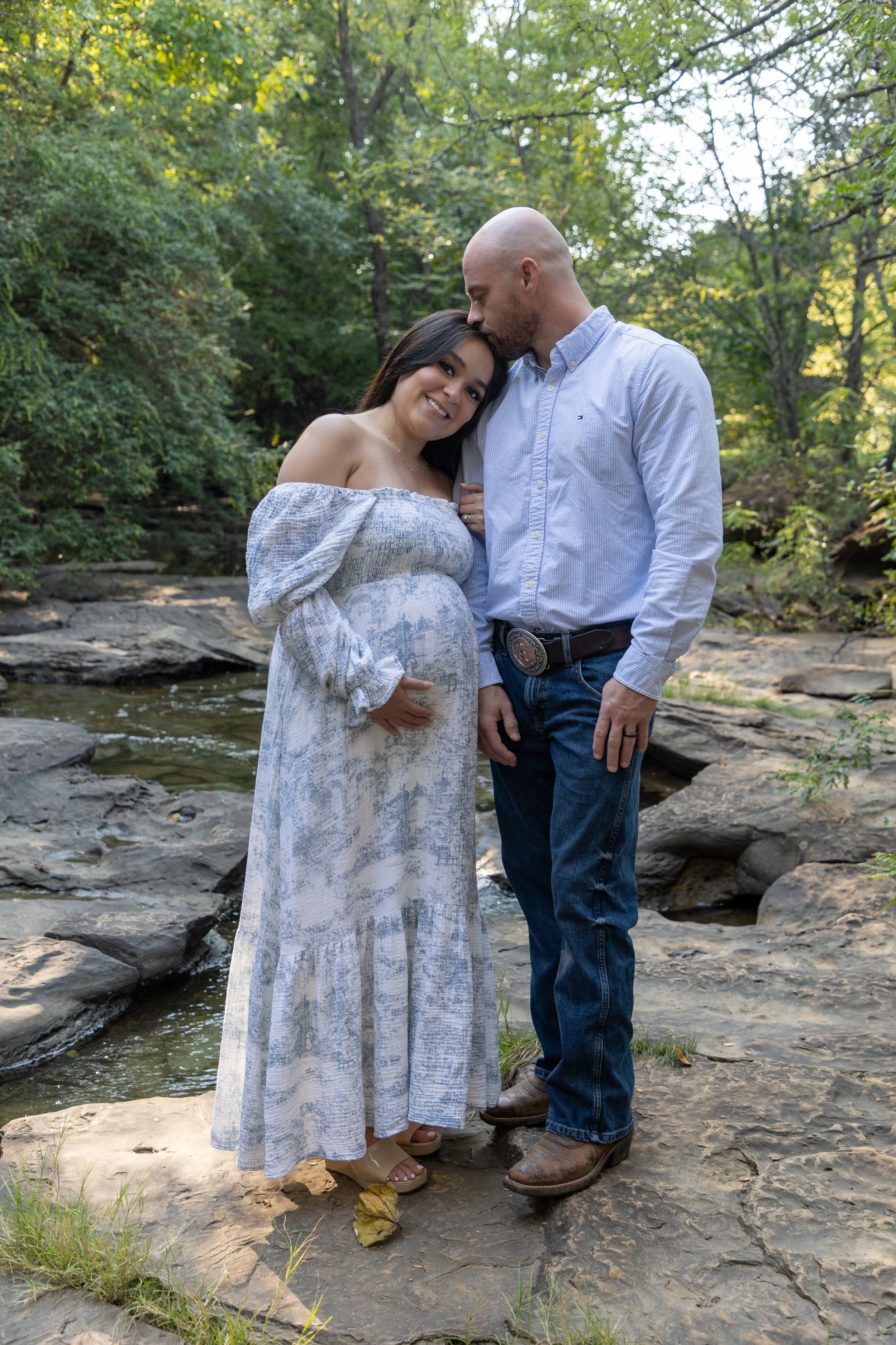 Pregnant woman and man standing on rocks by a creek in a wooded area, with trees and sunlight in the background.