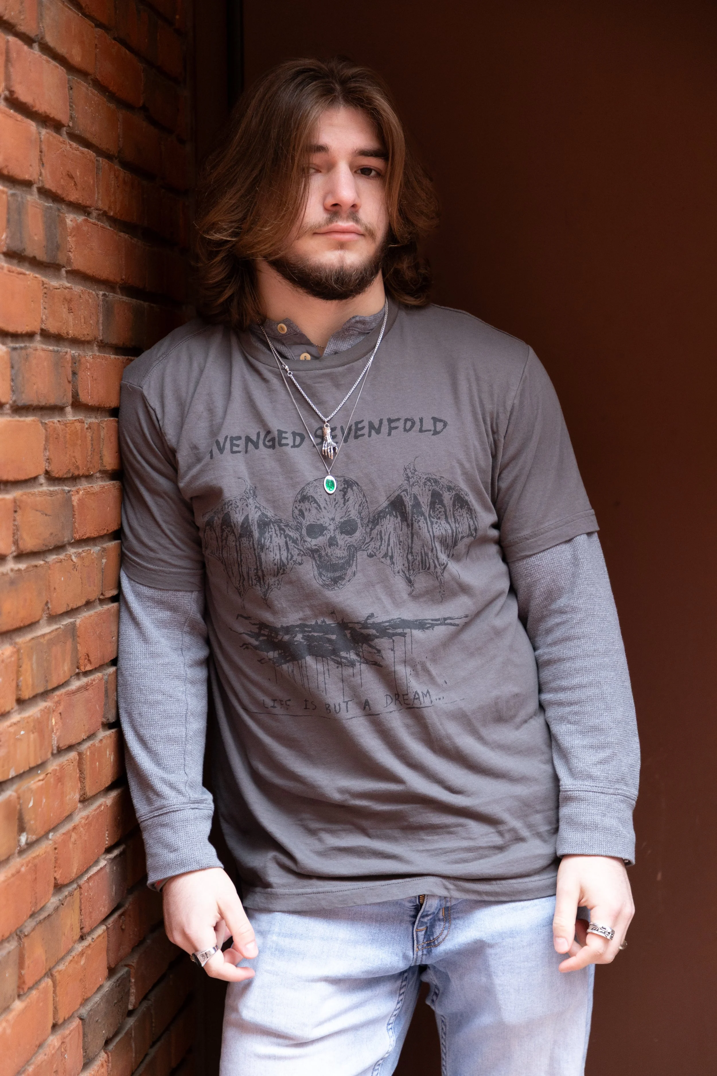 A young man with long brown hair, beard, wearing a gray Avenged Sevenfold t-shirt, layered over a long sleeve shirt, stands against a brick wall in an alleyway.