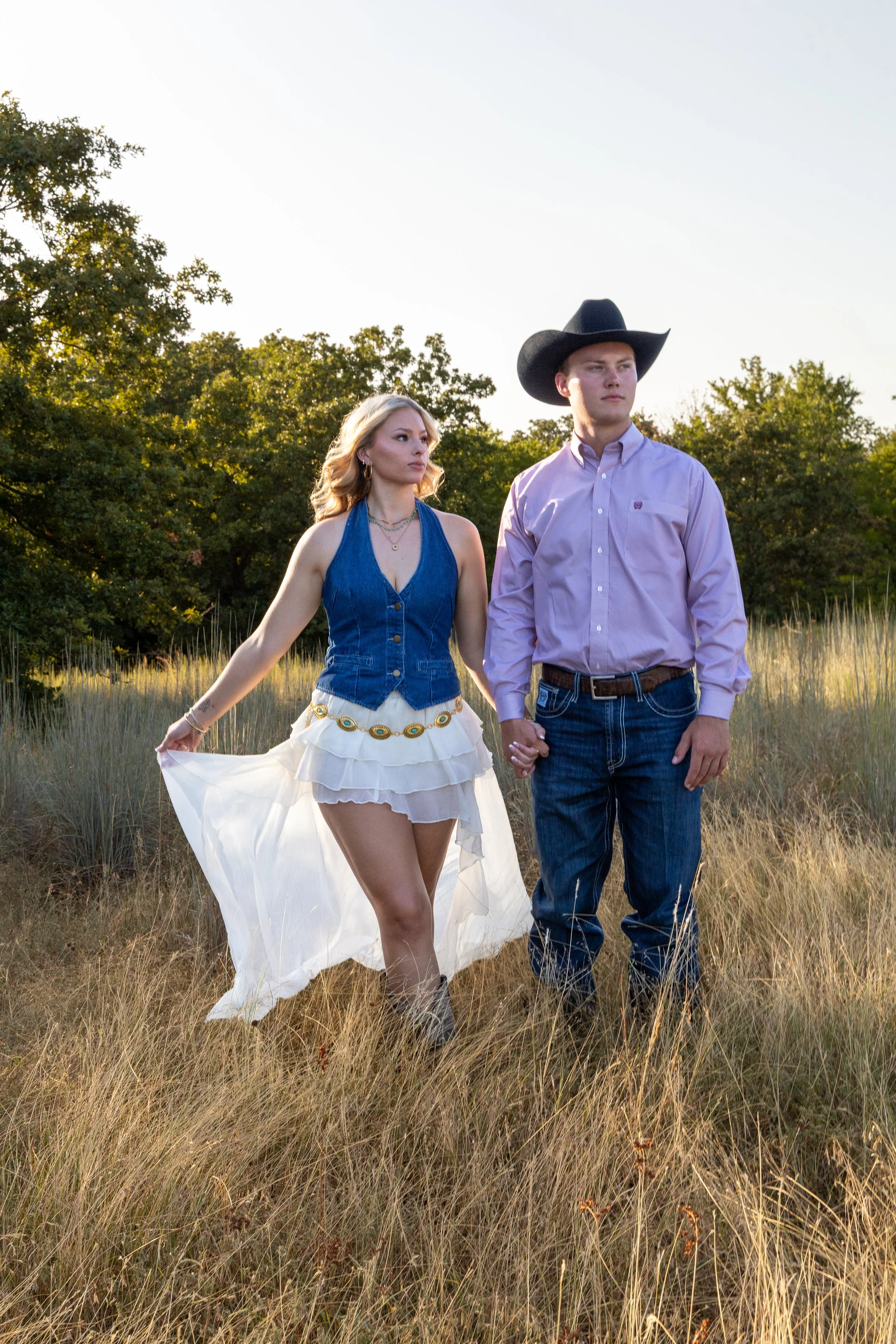 A young woman in a white flowing skirt and denim vest holding hands with a young man in a pink button-up shirt and cowboy hat, standing in a grassy field with trees in the background during sunset.