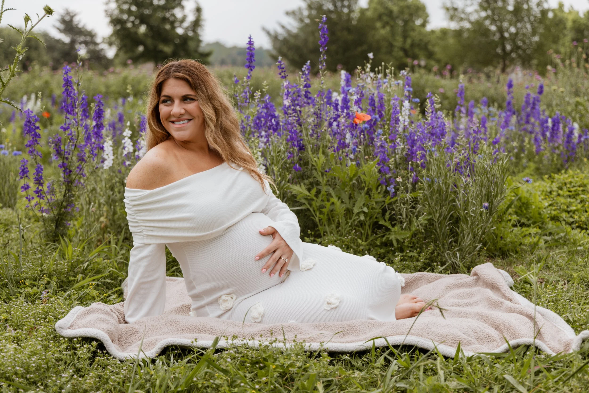 A pregnant woman with long, wavy hair, smiling and dressed in a white off-shoulder dress, sitting on a blanket in a field of purple and white flowers, with trees in the background.