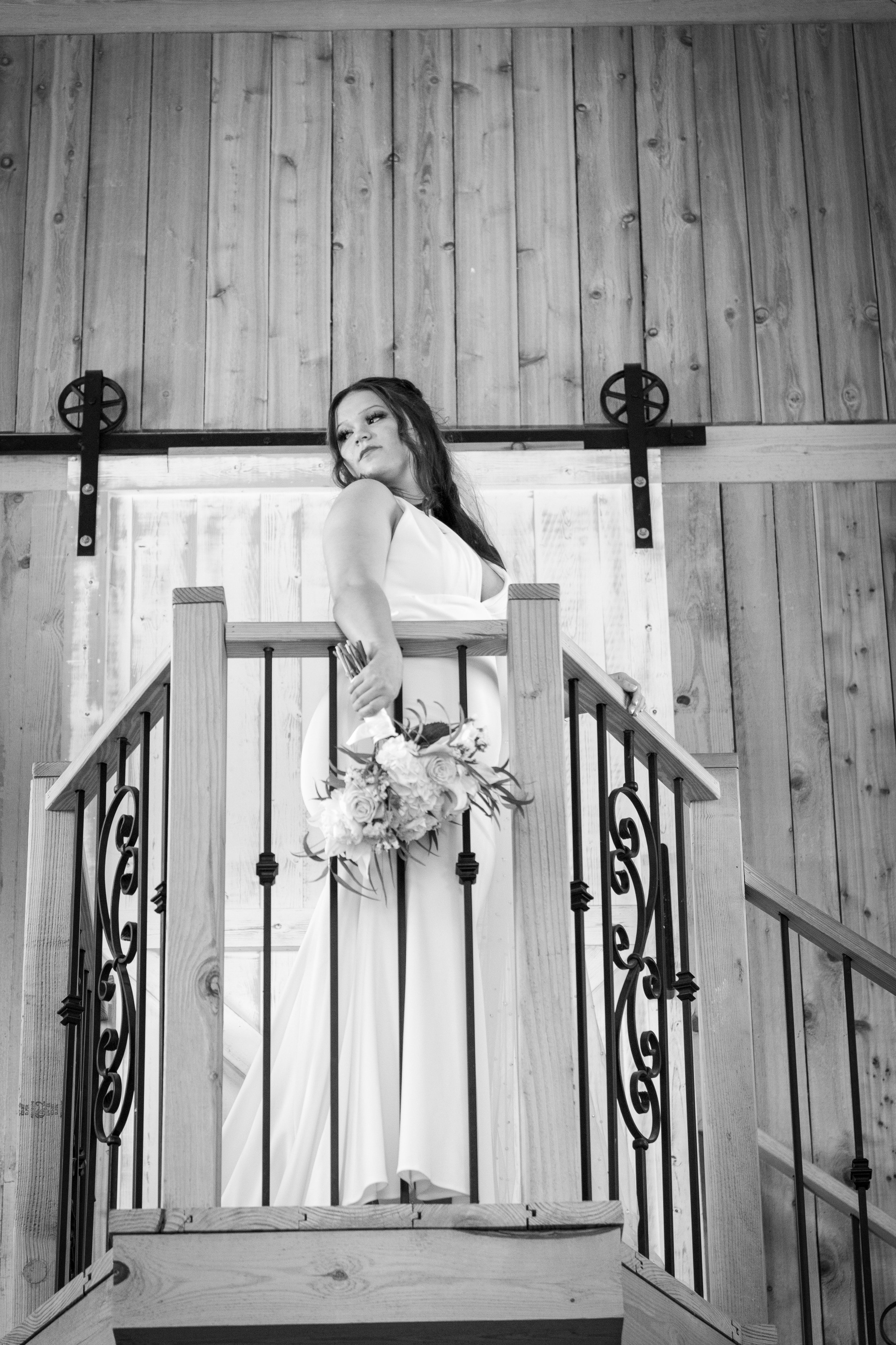 A woman in a white dress standing on a wooden staircase, holding a bouquet of flowers, with a wood-paneled wall background.