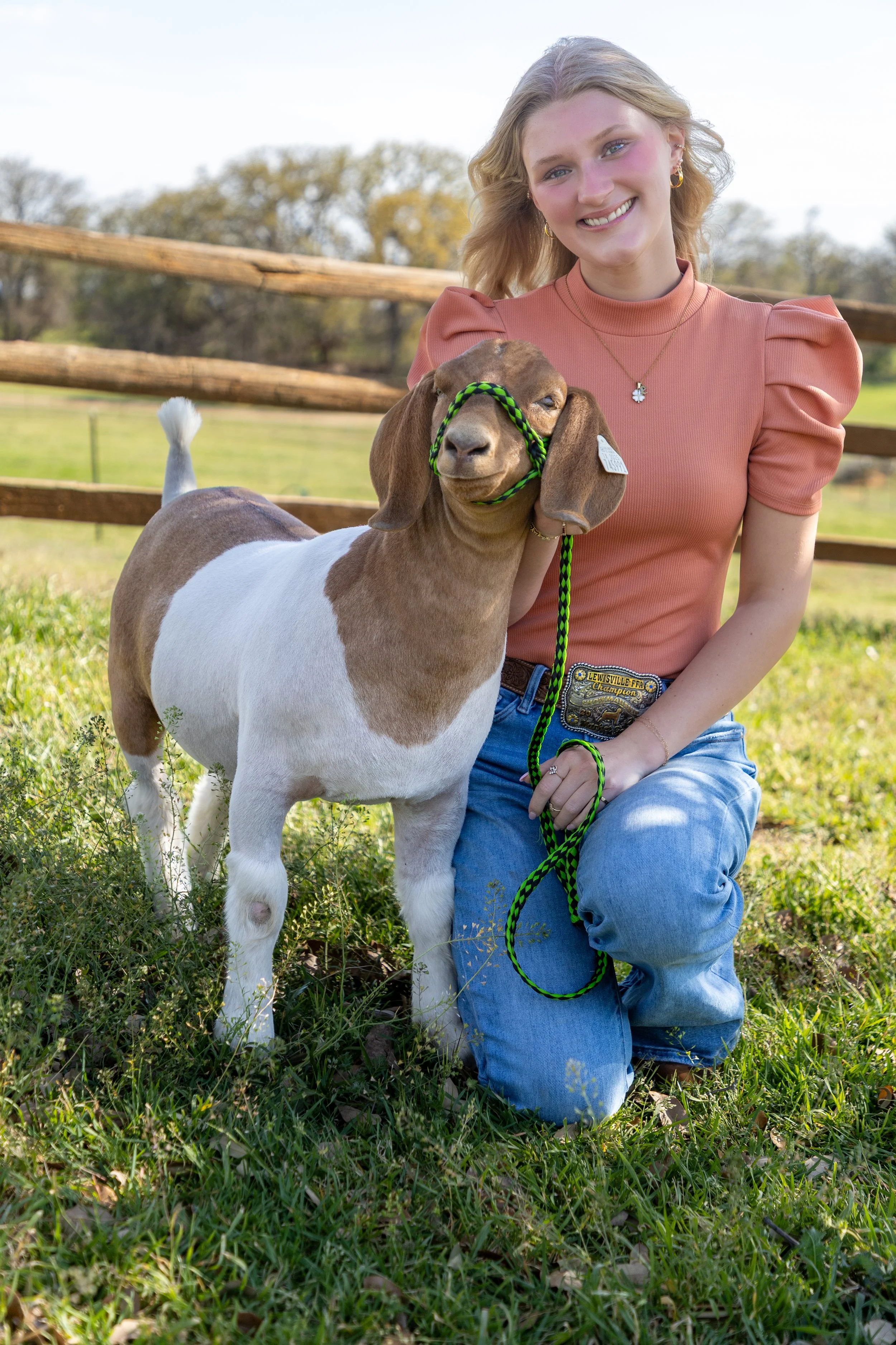 A young woman kneeling on grass with a goat on a leash, smiling outdoors in a farm or countryside setting.