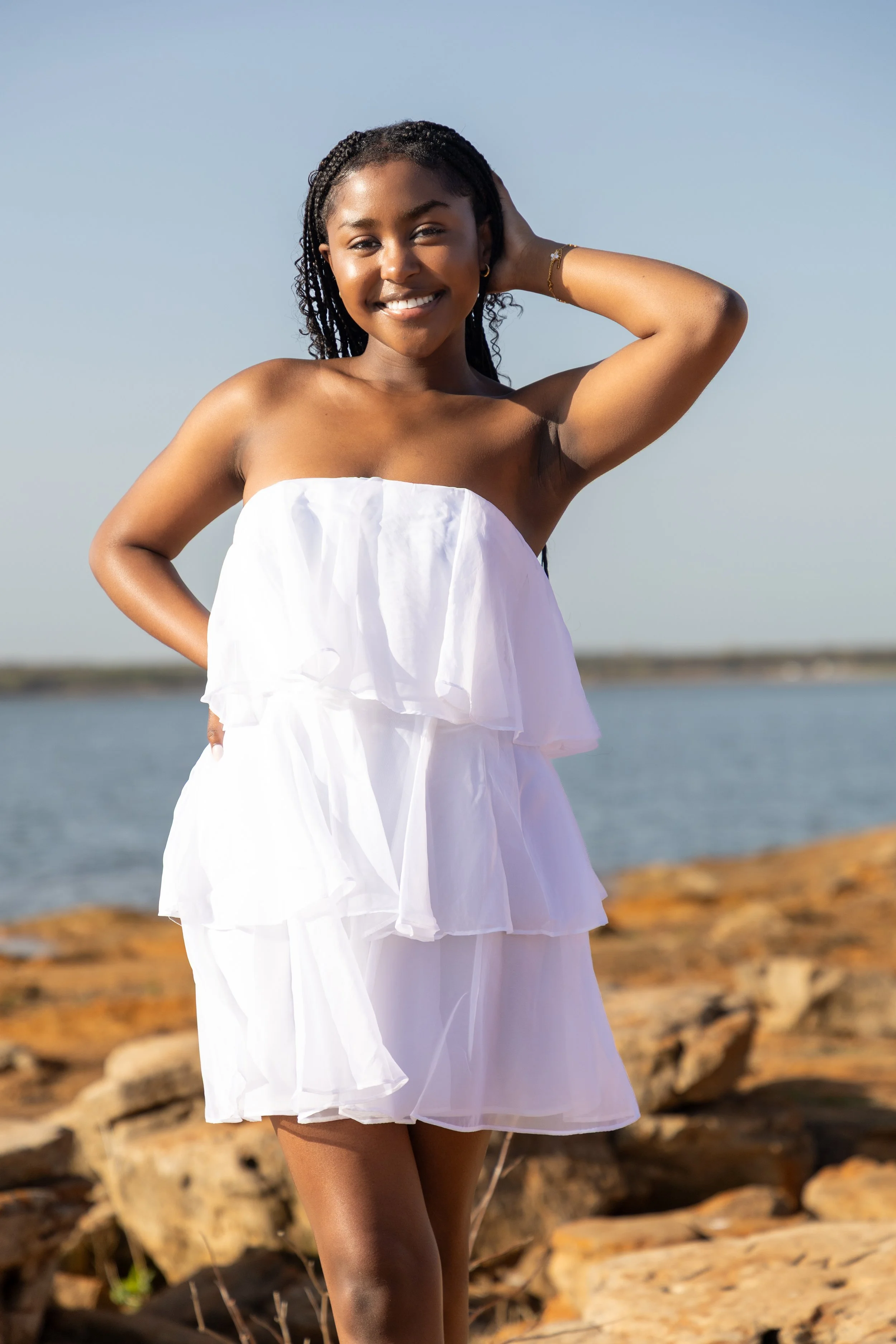 Young woman in a white strapless dress standing on rocky shoreline near water, smiling at the camera.