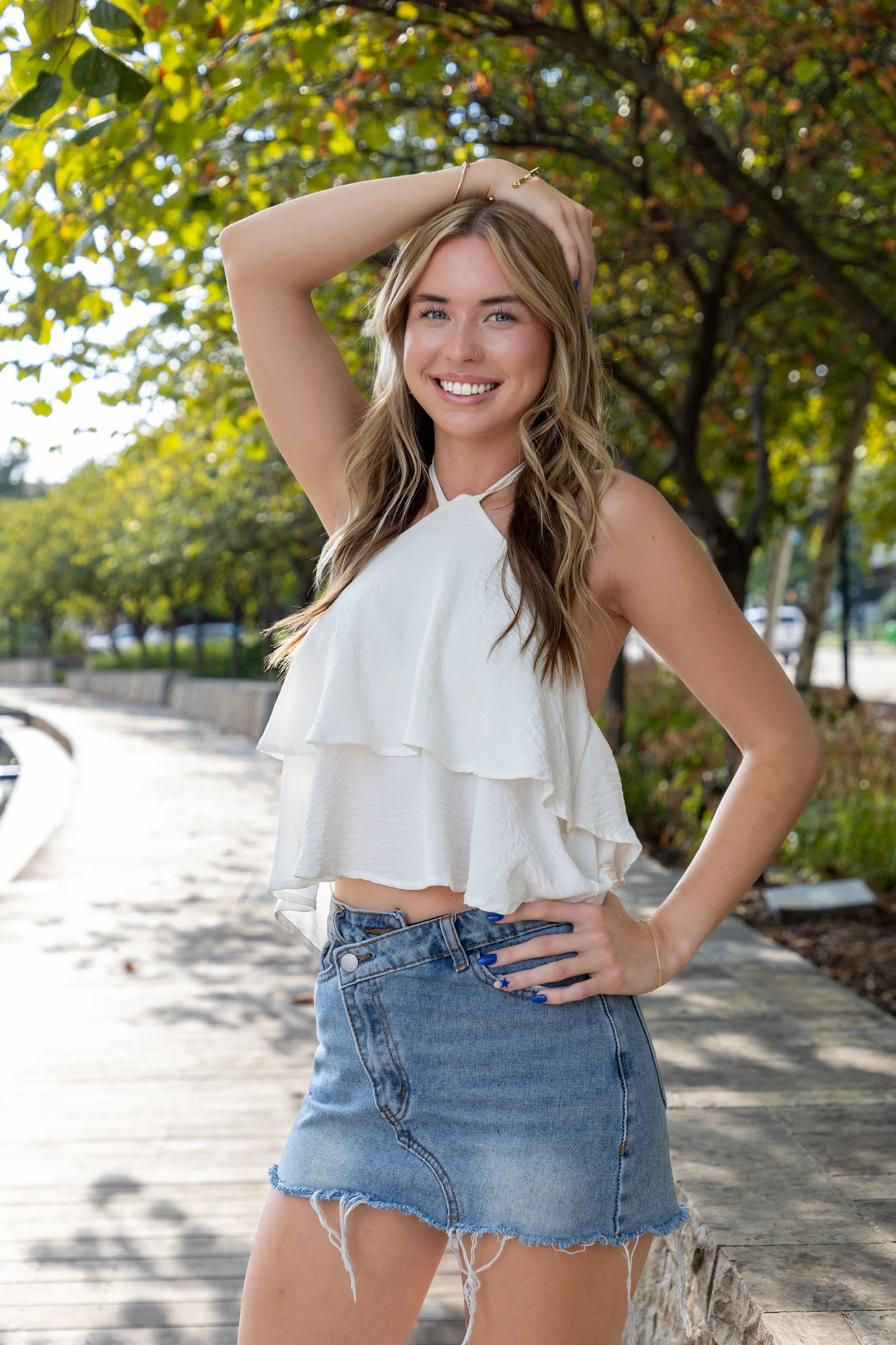 A smiling young woman with blonde hair, wearing a white sleeveless top and a denim skirt, posing outdoors on a sunny day among green trees.