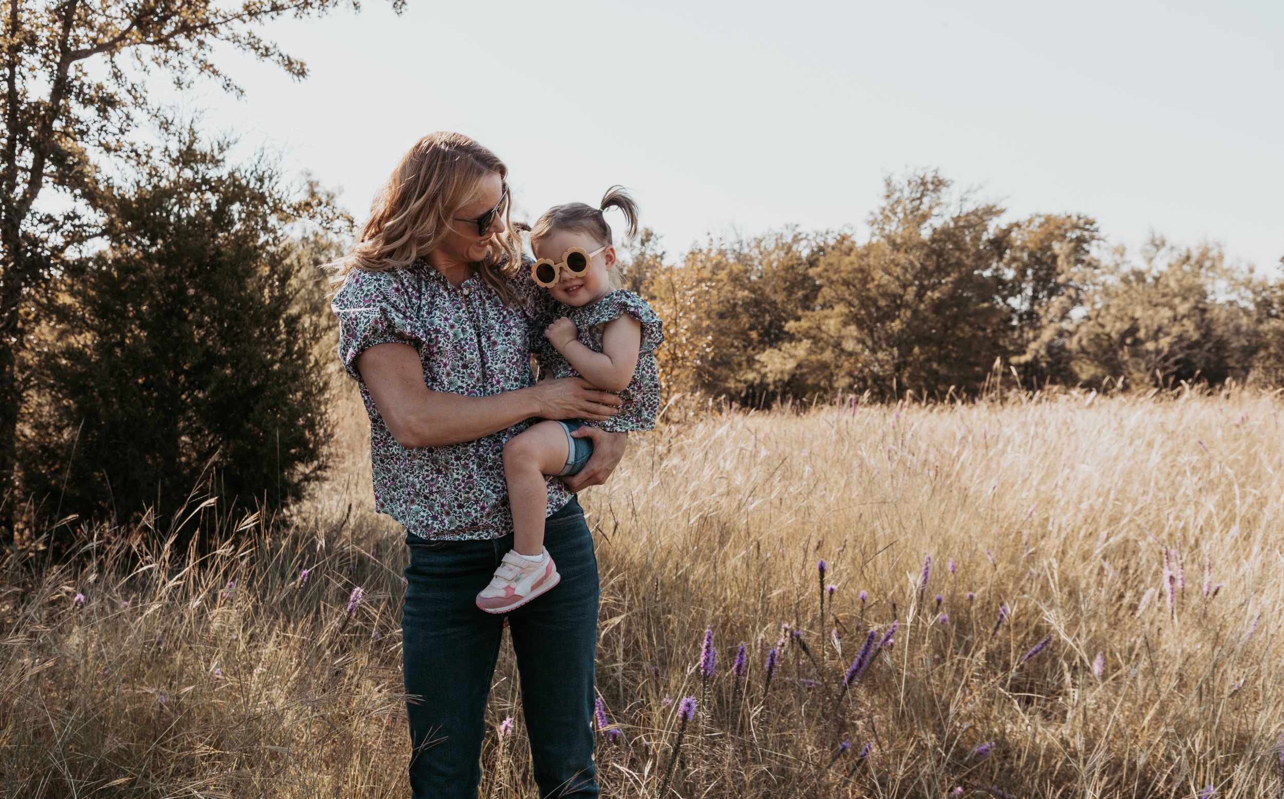 A woman holding a girl in a field with tall grass and purple flowers, during sunset or late afternoon, with trees in the background.