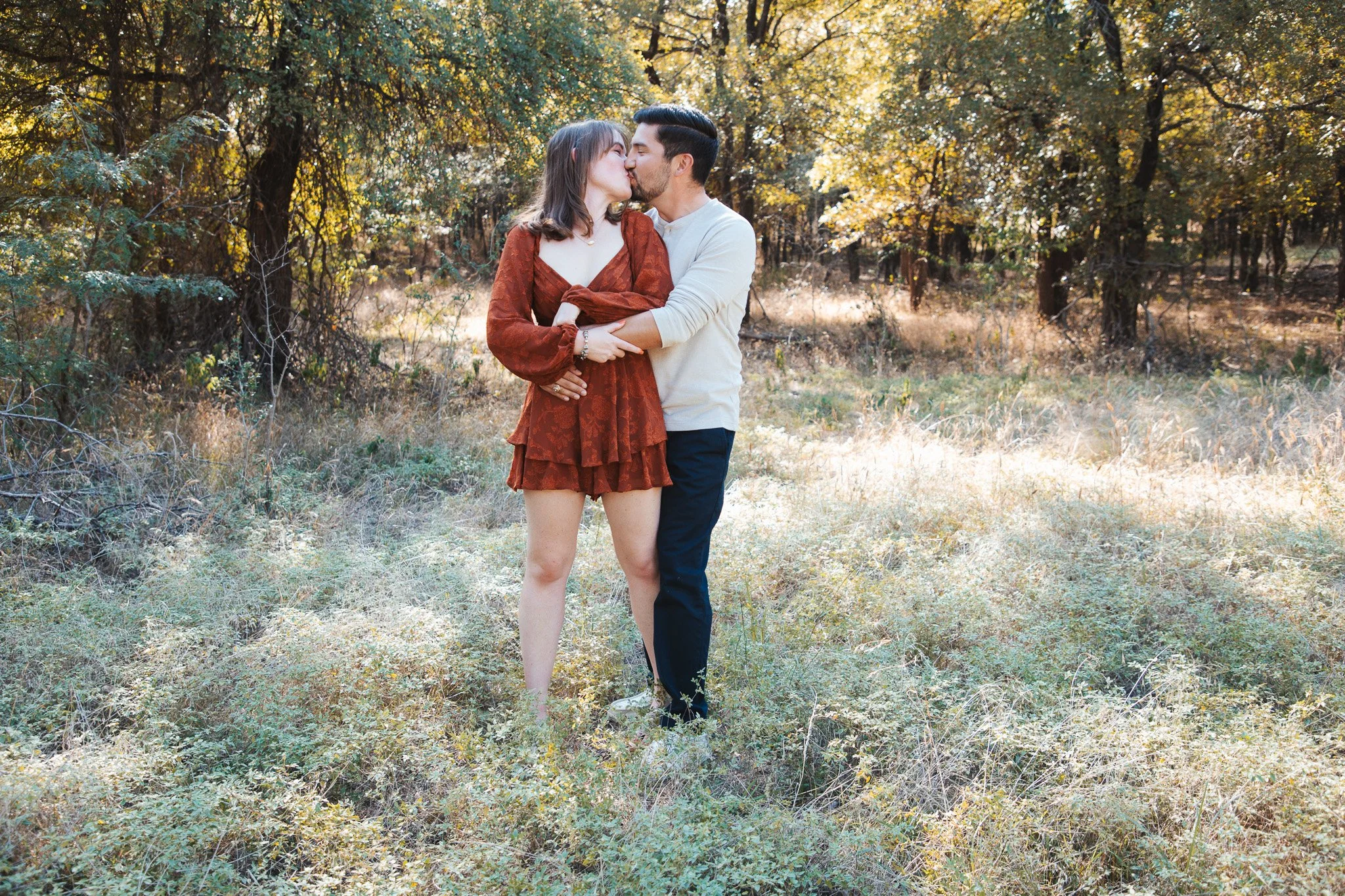 A couple sharing a kiss in a field during autumn, surrounded by trees with fall foliage.