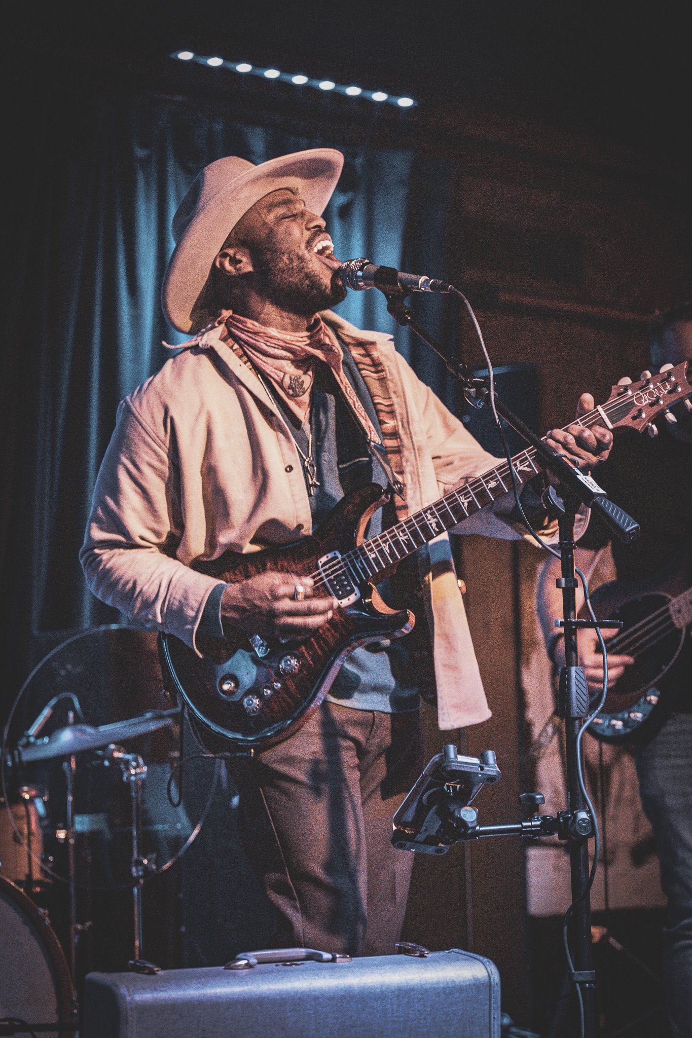 A male musician wearing a wide-brimmed hat and a neck scarf playing an electric guitar and singing into a microphone during a live performance.