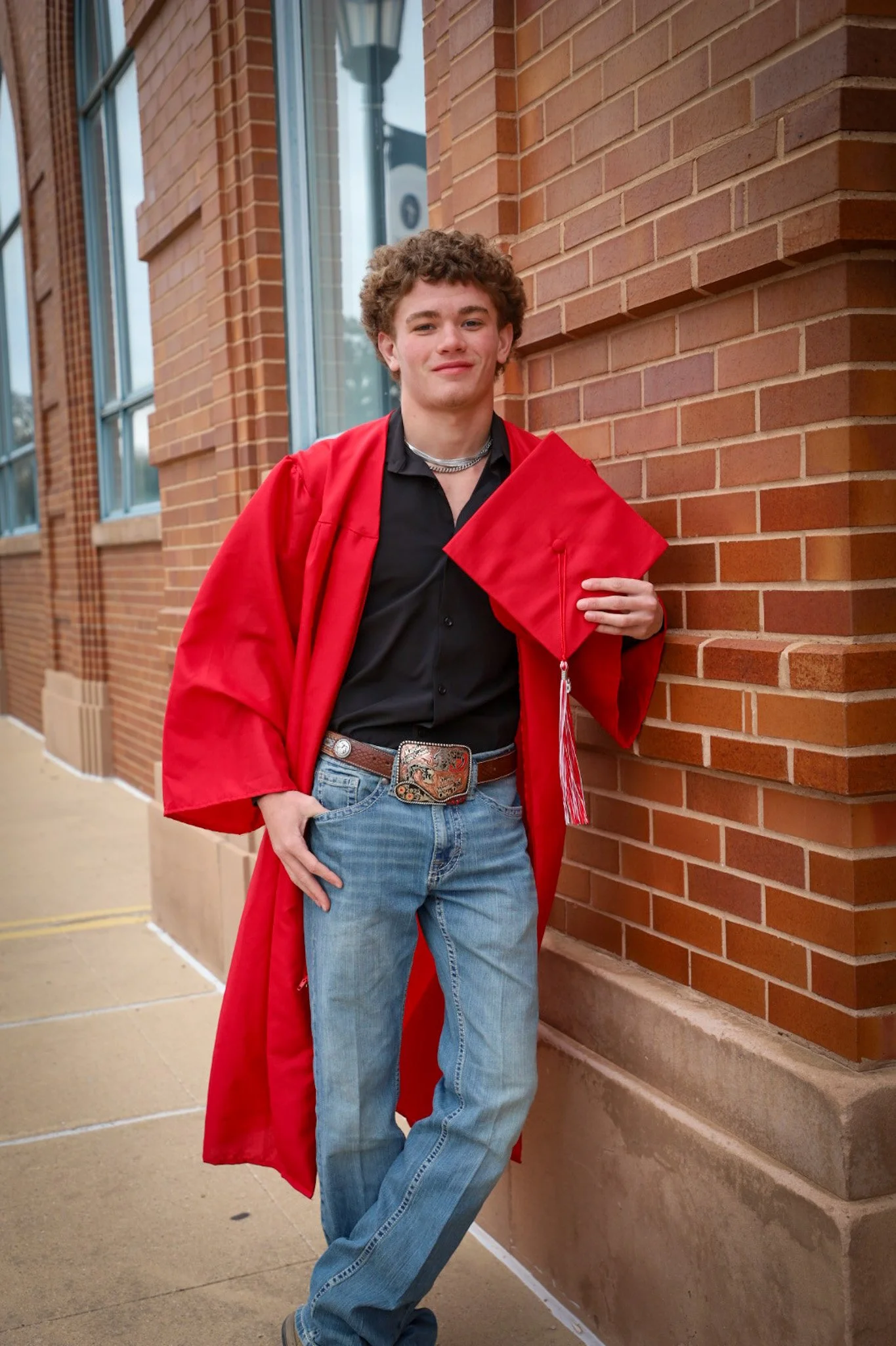 A young man in a red graduation gown and cap leaning against a brick wall, holding his cap in his right hand, outdoors.
