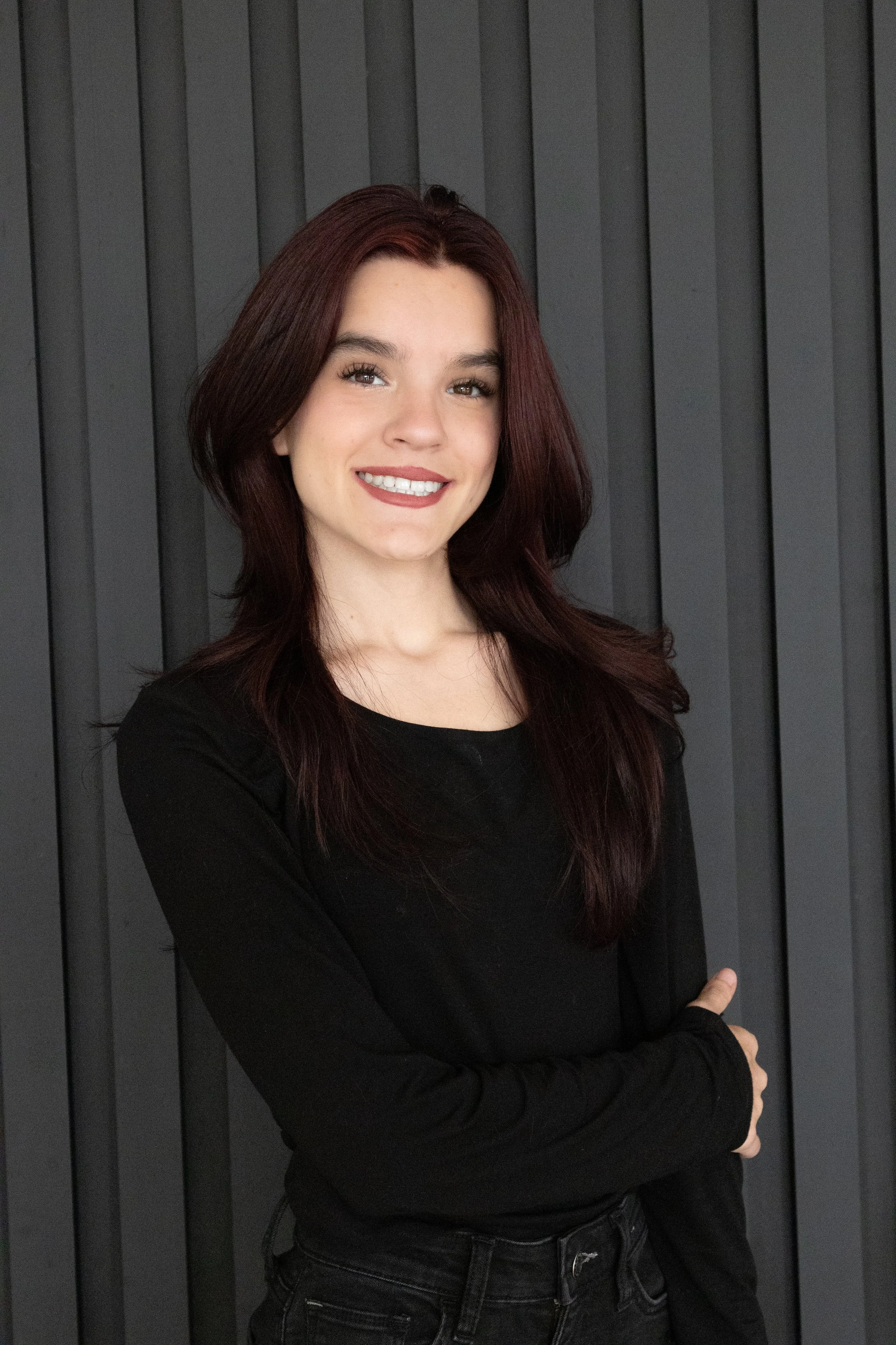 A young woman with long, dark red hair and a bright smile, standing with her arms crossed in front of a dark gray paneled wall, wearing a black long-sleeve top.