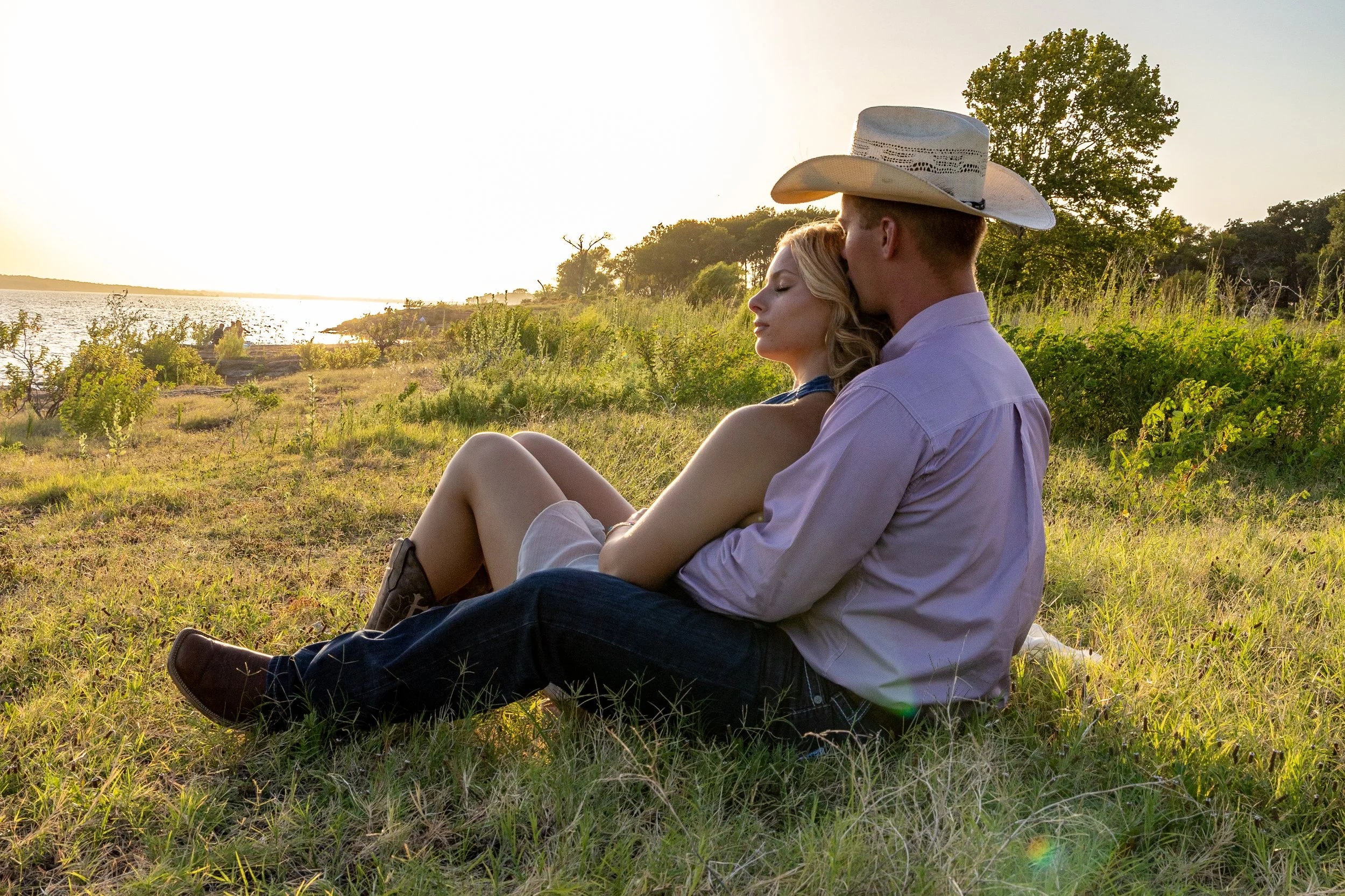 A couple sitting on grass near a body of water during sunset, the man wearing a cowboy hat and the woman with closed eyes resting her head on his shoulder.