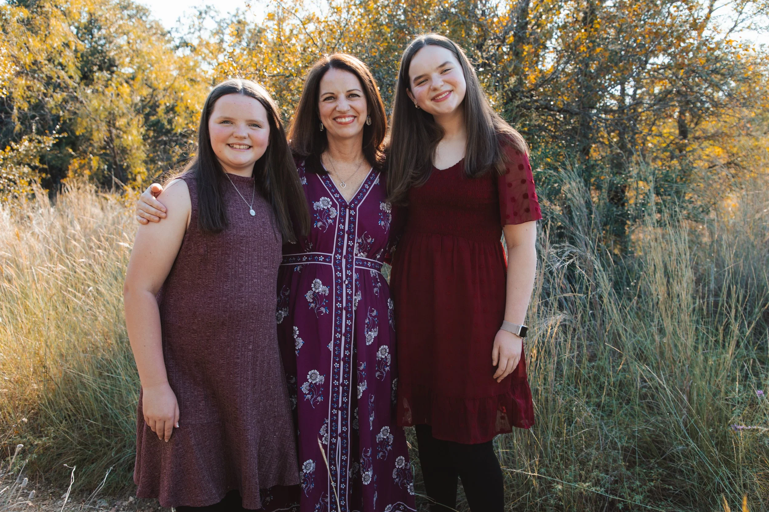 A woman with two young girls standing outdoors in a grassy area, smiling, with trees and autumn foliage in the background.