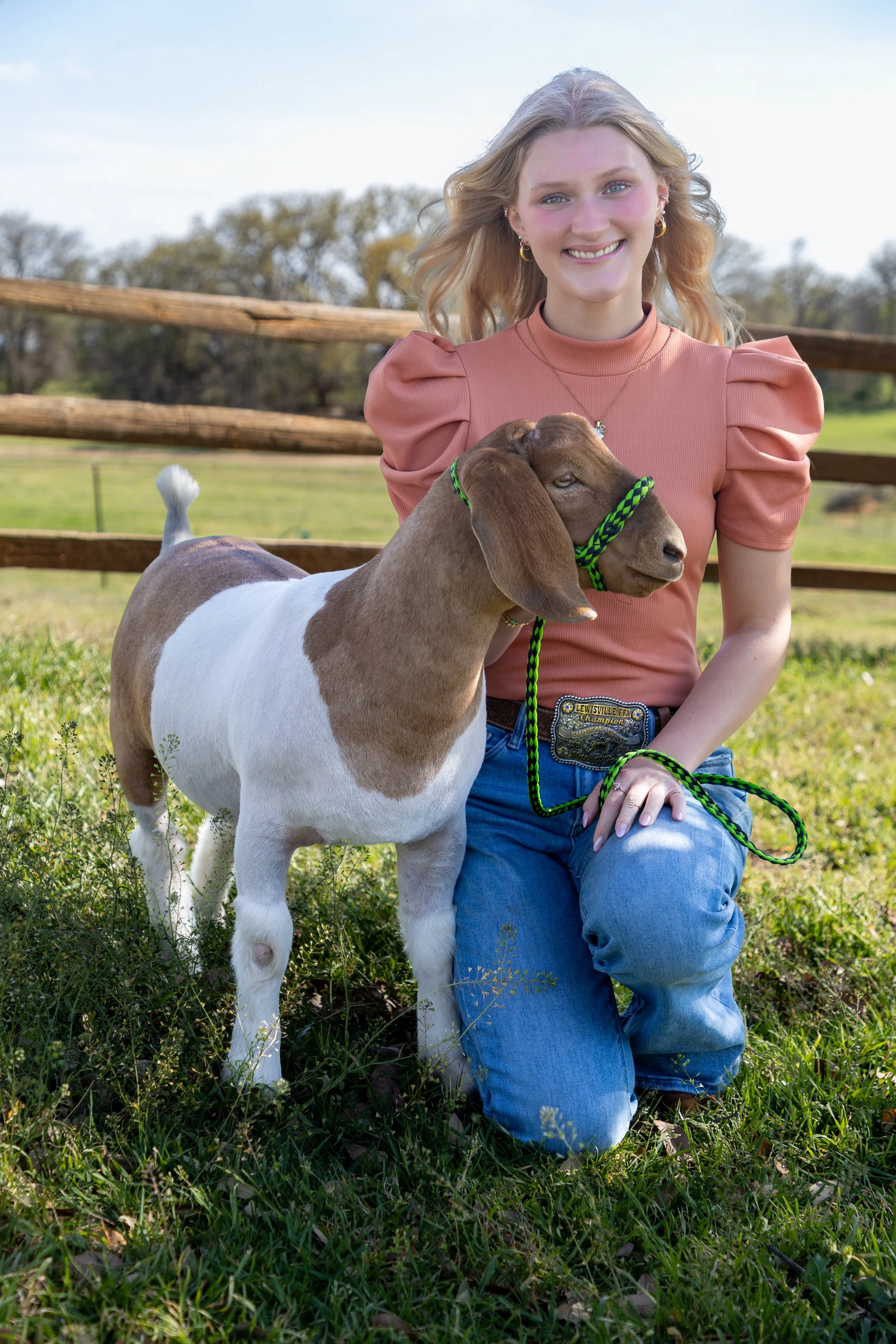 A young woman with blonde hair kneeling on the grass in a field, smiling at the camera, holding a brown and white goat with long ears on a green leash. She is wearing a pink top with puffed sleeves, blue jeans, and a large belt buckle. There is a woo