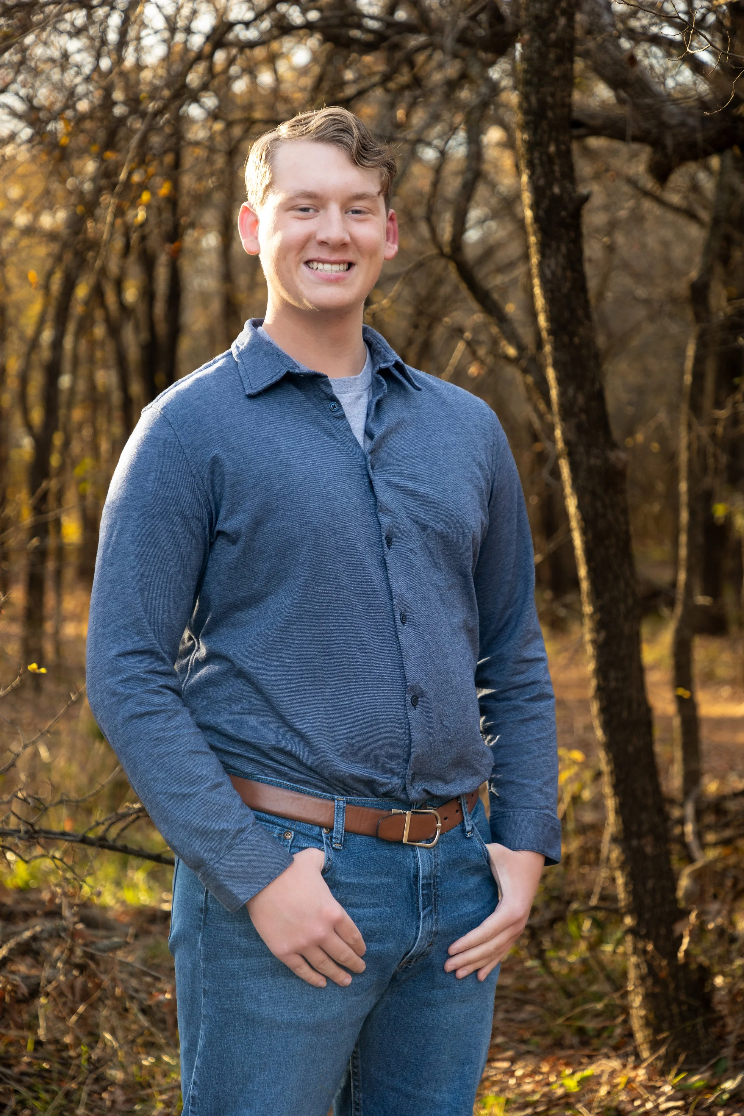 A young man smiling outdoors in autumn, wearing a blue button-up shirt and jeans, standing in a wooded area with trees and fallen leaves.