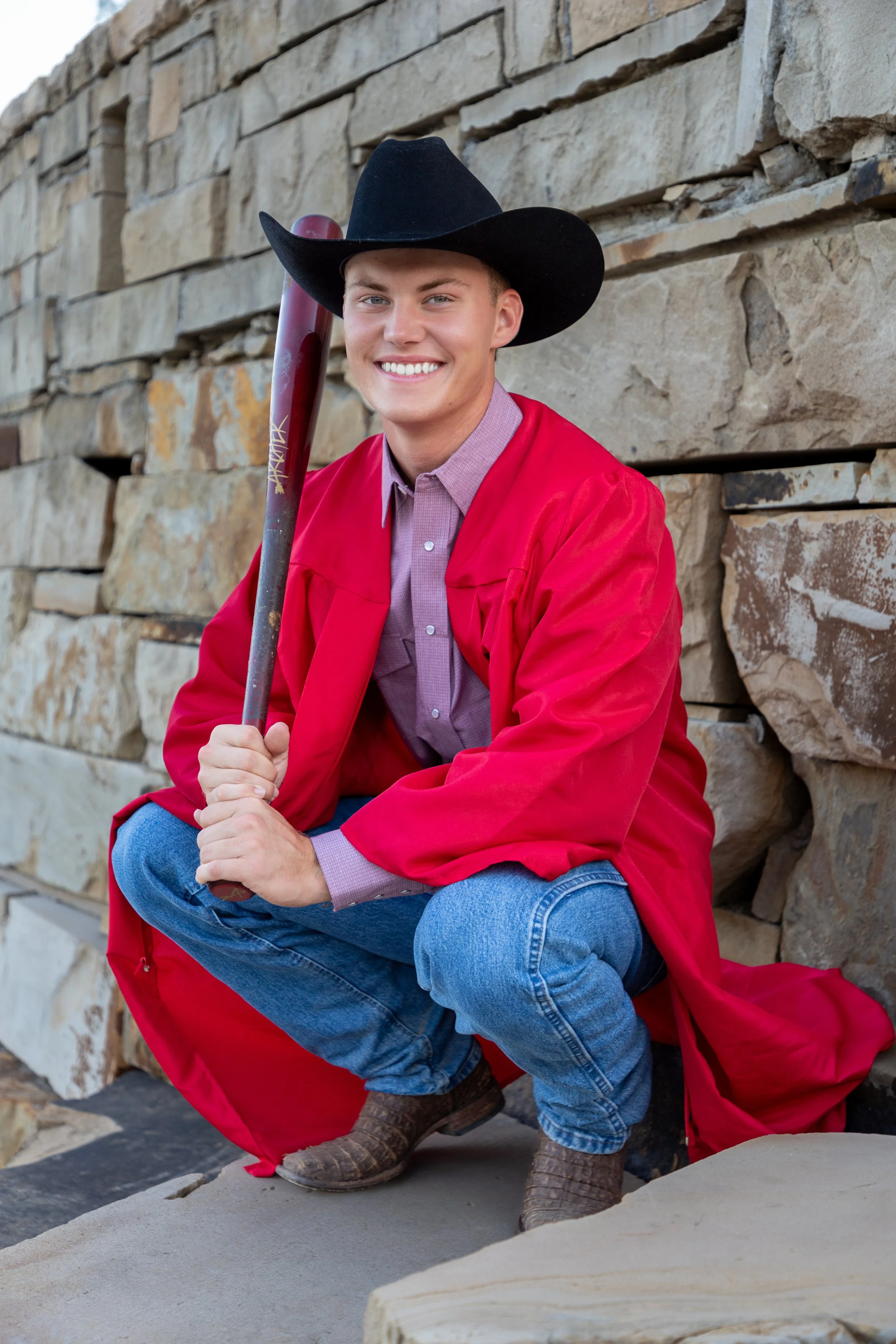 Young man dressed in a red graduation gown and cowboy hat, squatting with a baseball bat, smiling in front of a stone wall.
