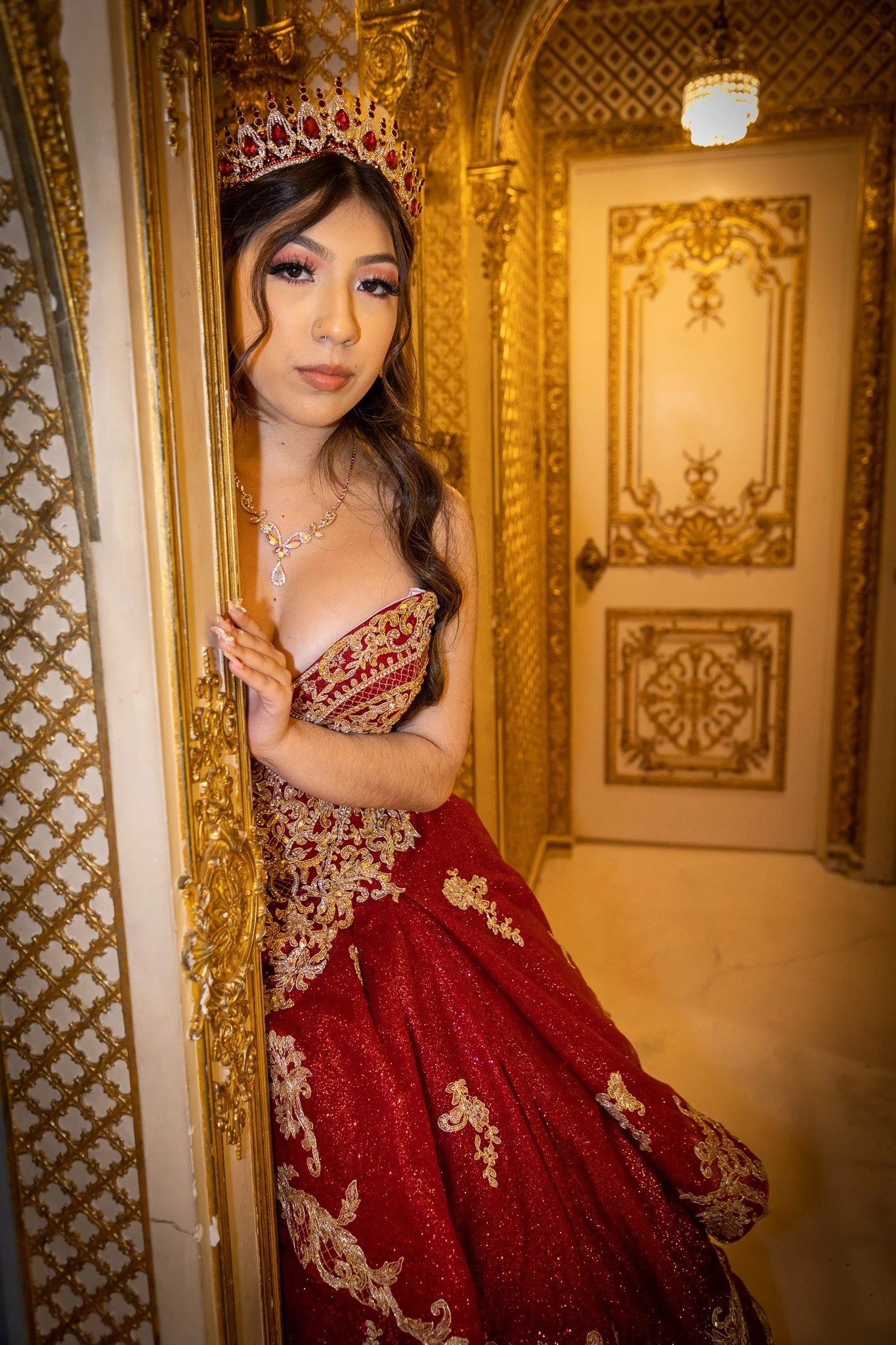 A young woman in an ornate red and gold gown and crown, peeking out from behind a decorated gold wall, in a lavishly decorated gold and white room.
