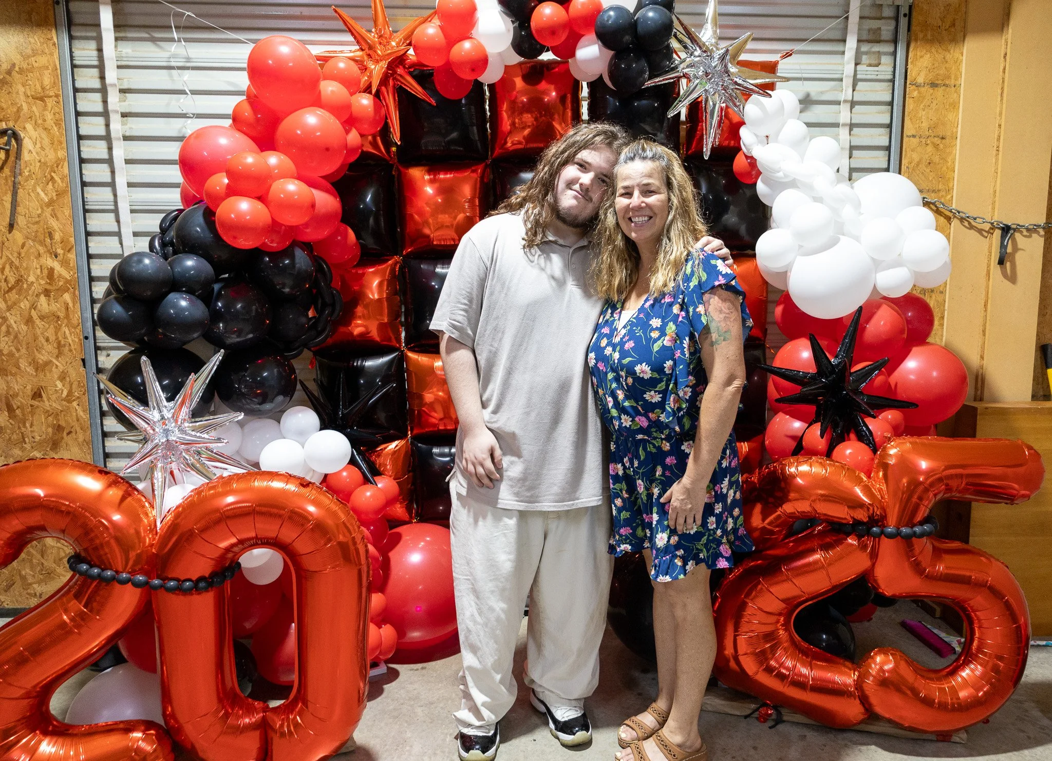 A young man and an older woman standing together in front of a colorful balloon backdrop with red, black, white, and silver balloons, star-shaped balloons, and large red foil balloons spelling '2023'. They are smiling and posing for the photo.