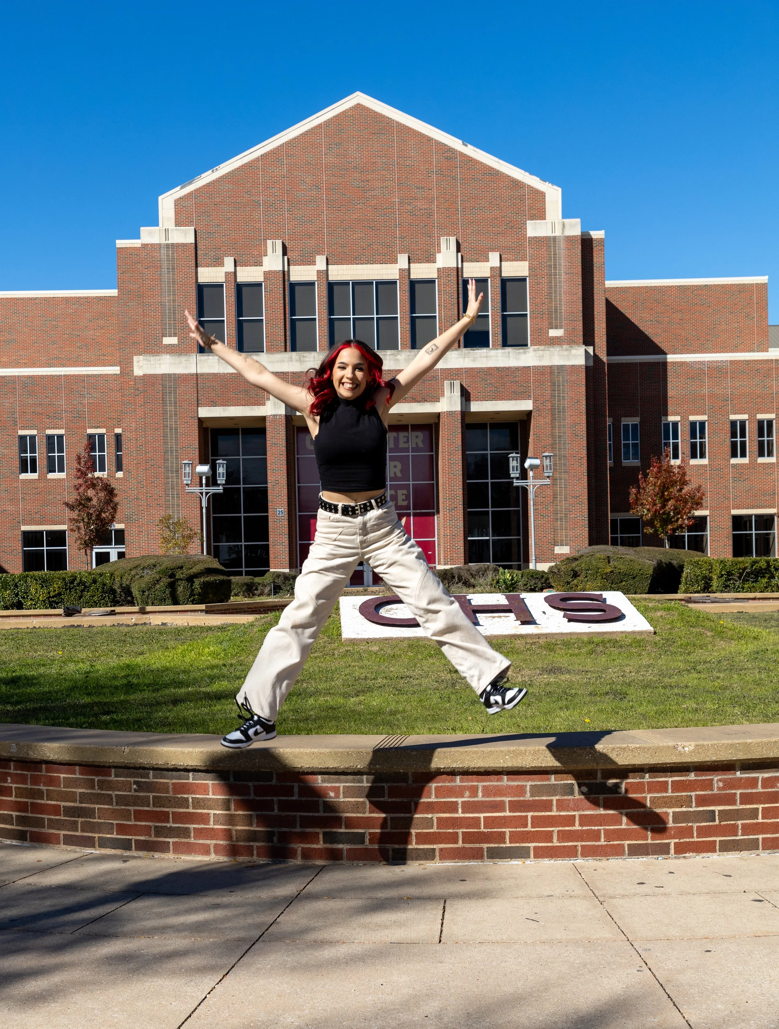 A young woman with red hair jumping in front of a brick school building on a sunny day.