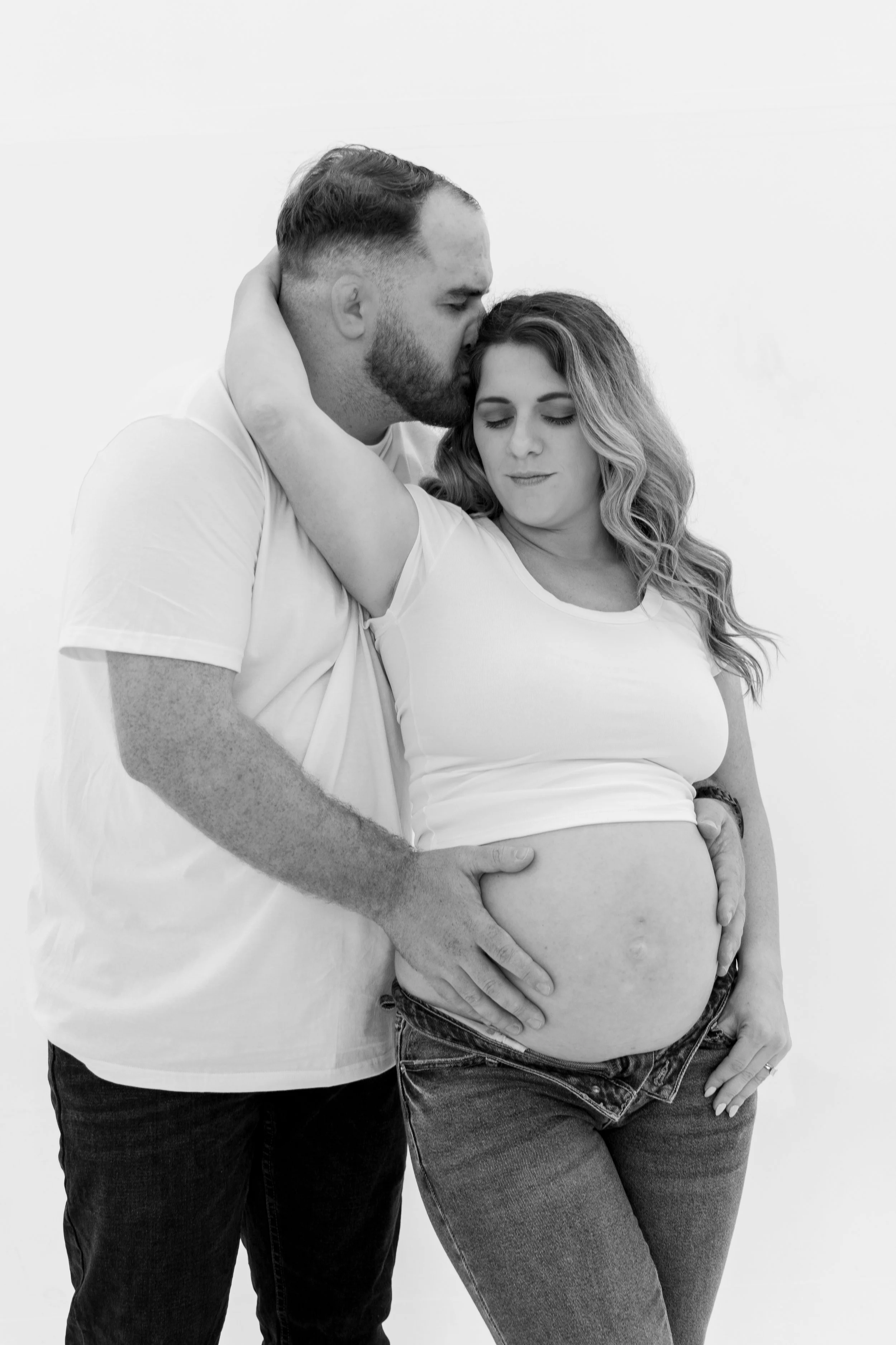 A pregnant woman with long wavy hair and wearing a white t-shirt and jeans, standing with her eyes closed, holding her belly, while a man with short hair, beard, and wearing a white t-shirt and black jeans, gently kisses her forehead and places his h