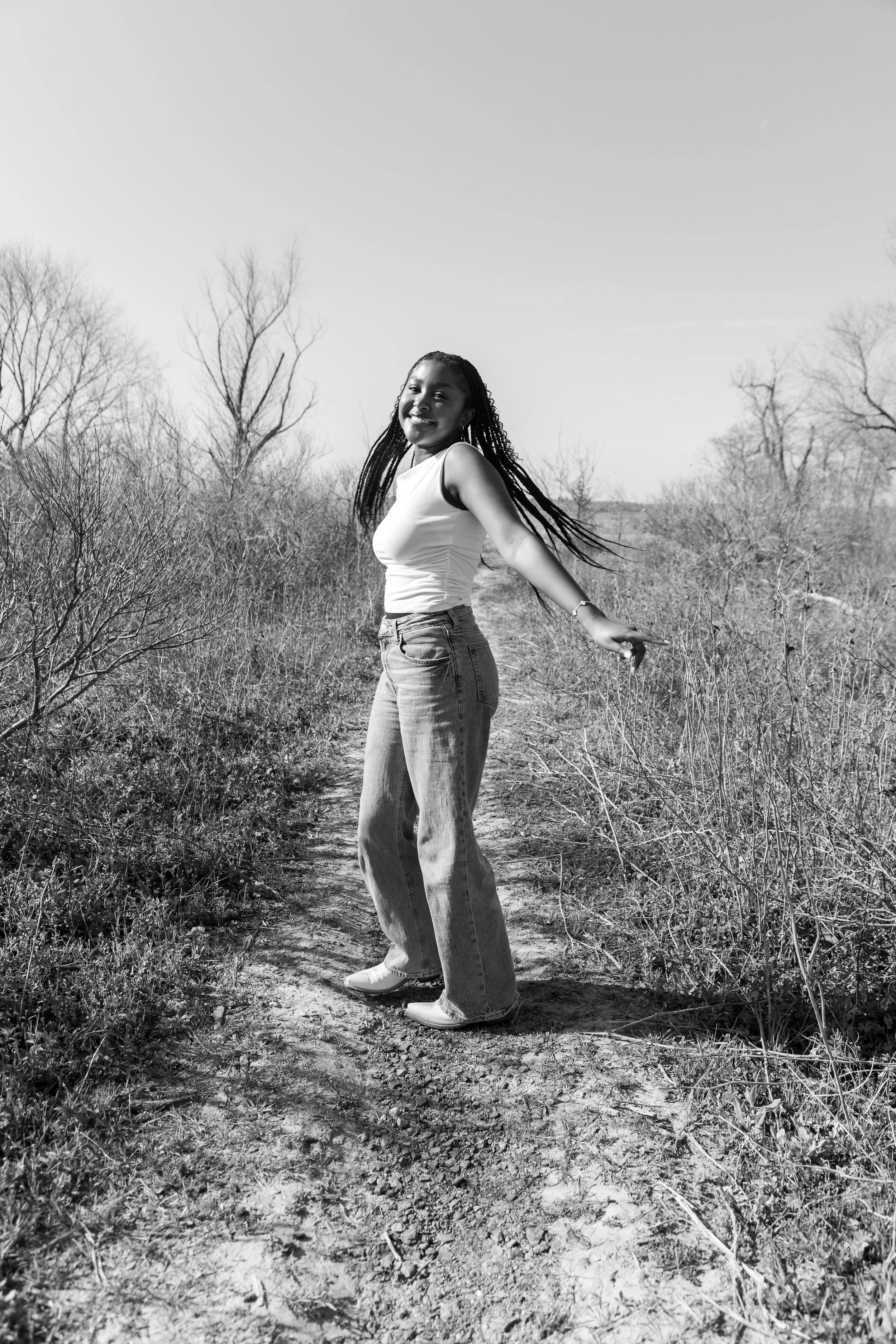 A woman with long braids smiling and dancing on a dirt trail in a natural, leafless landscape.