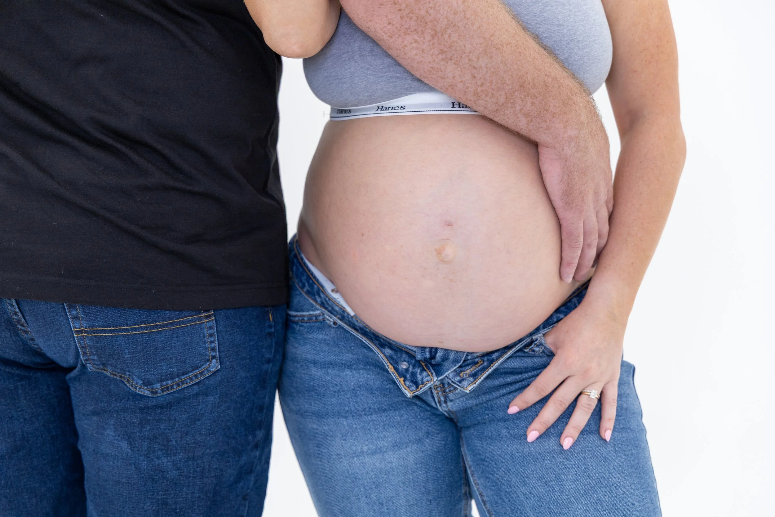 Close-up of a pregnant woman and a man standing close together, wearing jeans and casual tops, with the man's arm around her waist, highlighting her pregnant belly.