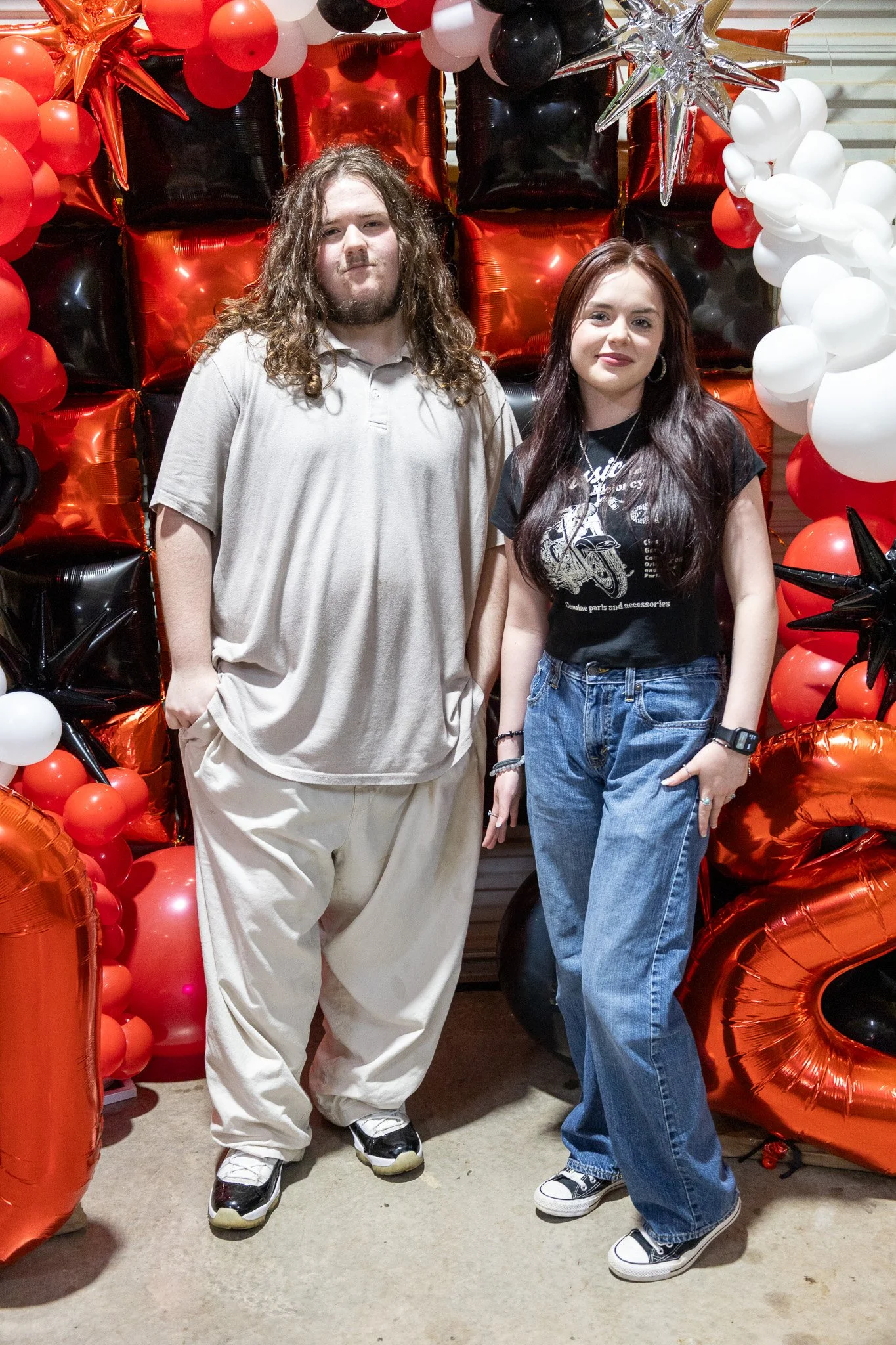 A man and woman standing together in front of a balloon backdrop with red, black, white, and orange balloons, some shaped like stars and numbers, at an indoor celebration or party.
