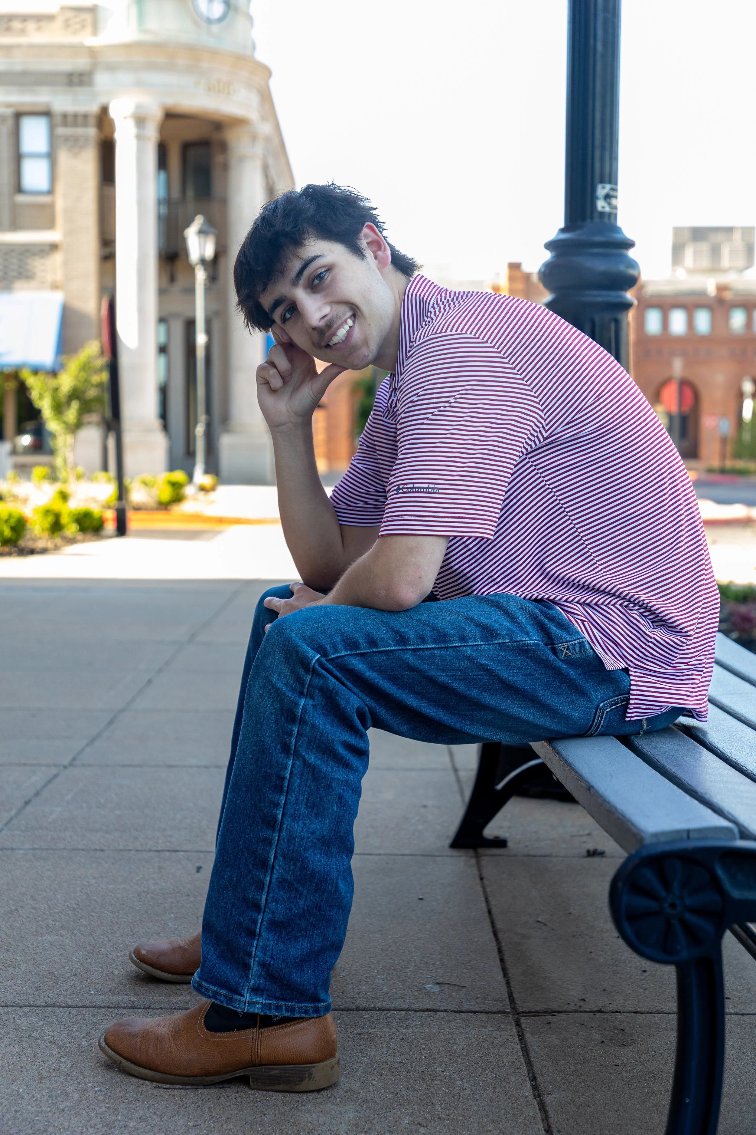 A young man sits on a park bench outdoors, smiling and leaning his head on his hand. He wears a striped polo shirt, blue jeans, and brown boots. In the background, there are historic buildings and a lamppost.