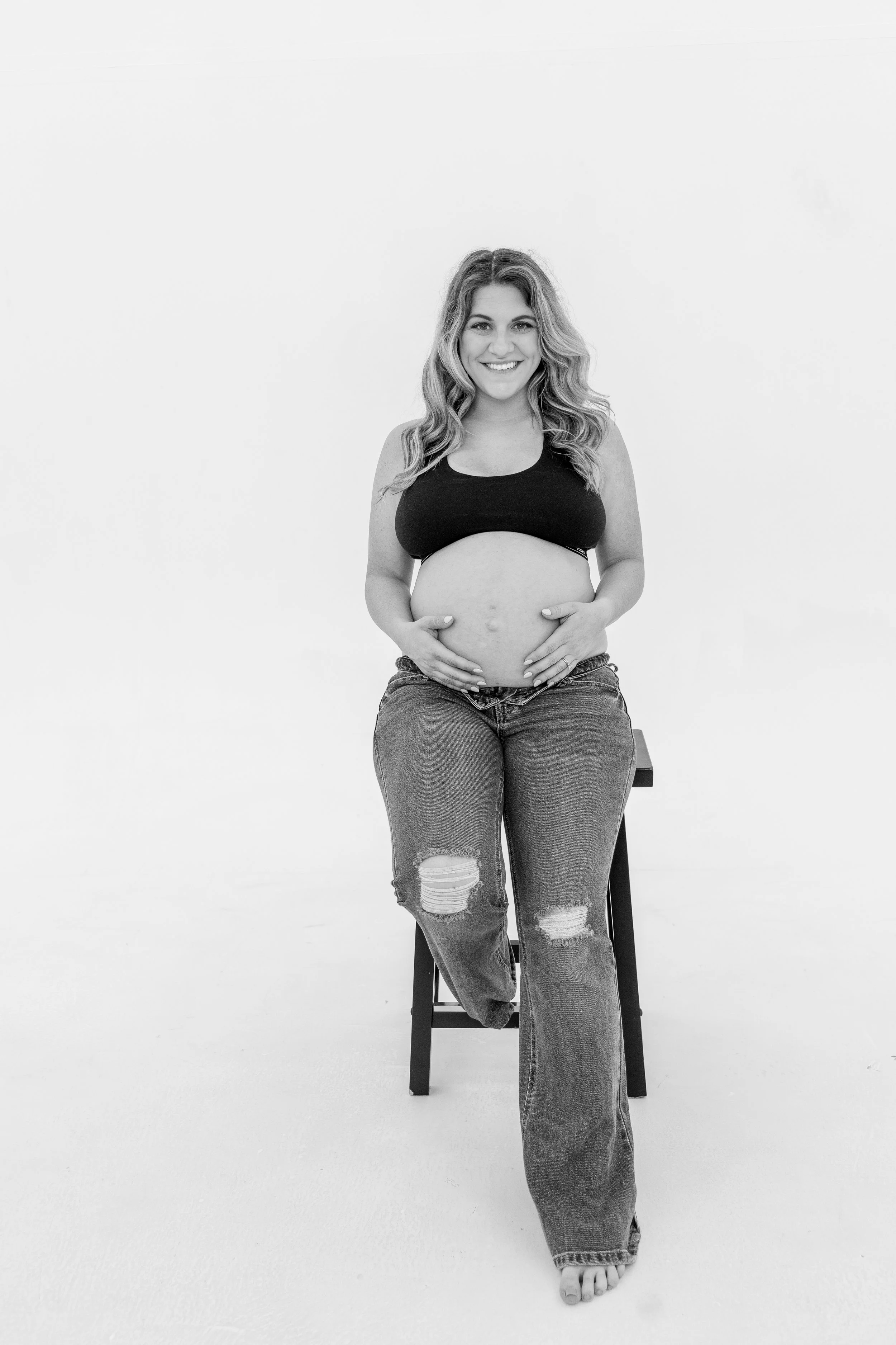 A pregnant woman sitting on a stool, smiling, wearing a black tank top and ripped jeans, with her hands on her belly, in a plain white studio.