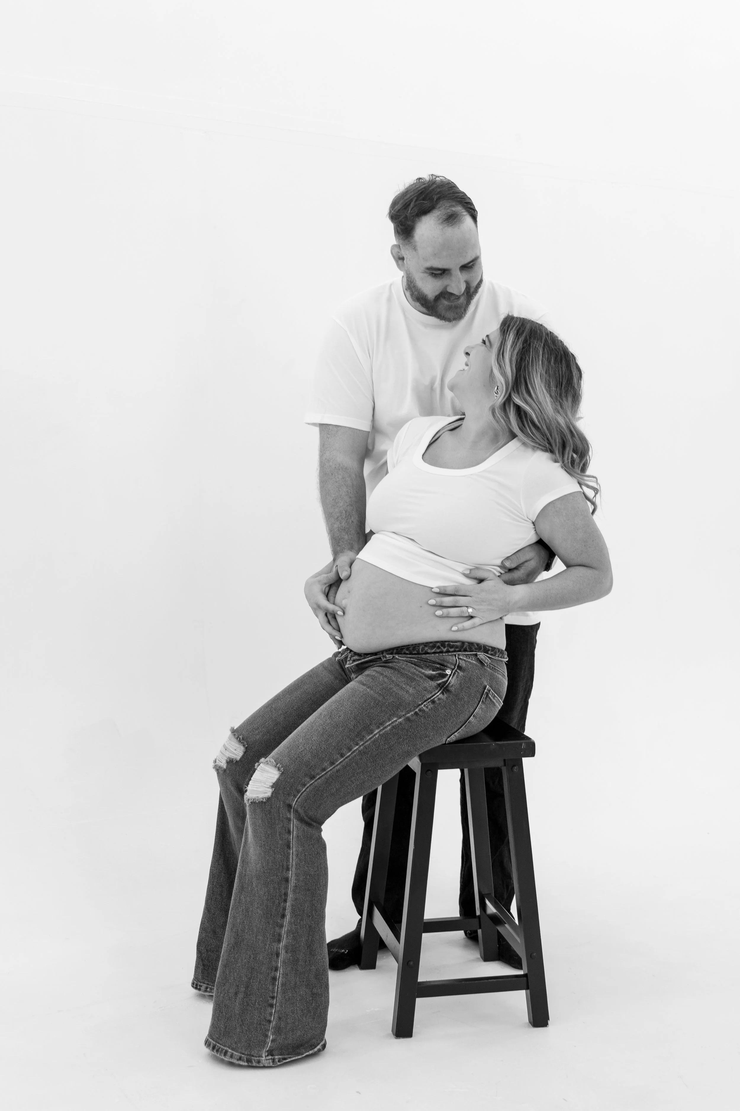 A pregnant woman with long hair sitting on a stool and smiling at a man who is standing and leaning over her with one hand on her belly, both are smiling and looking at each other in a minimalistic studio setting.