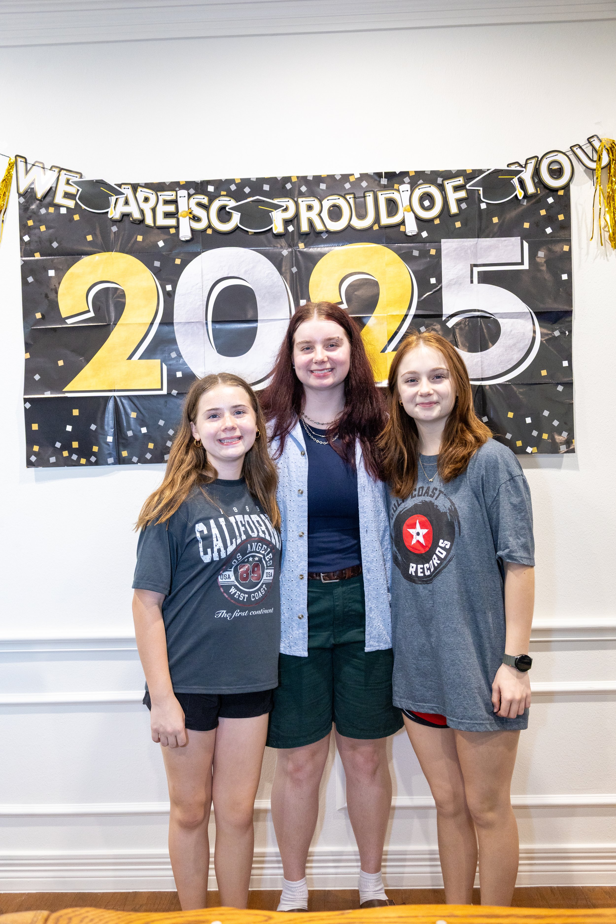 Three young women standing in front of a graduation celebration backdrop, smiling at the camera.