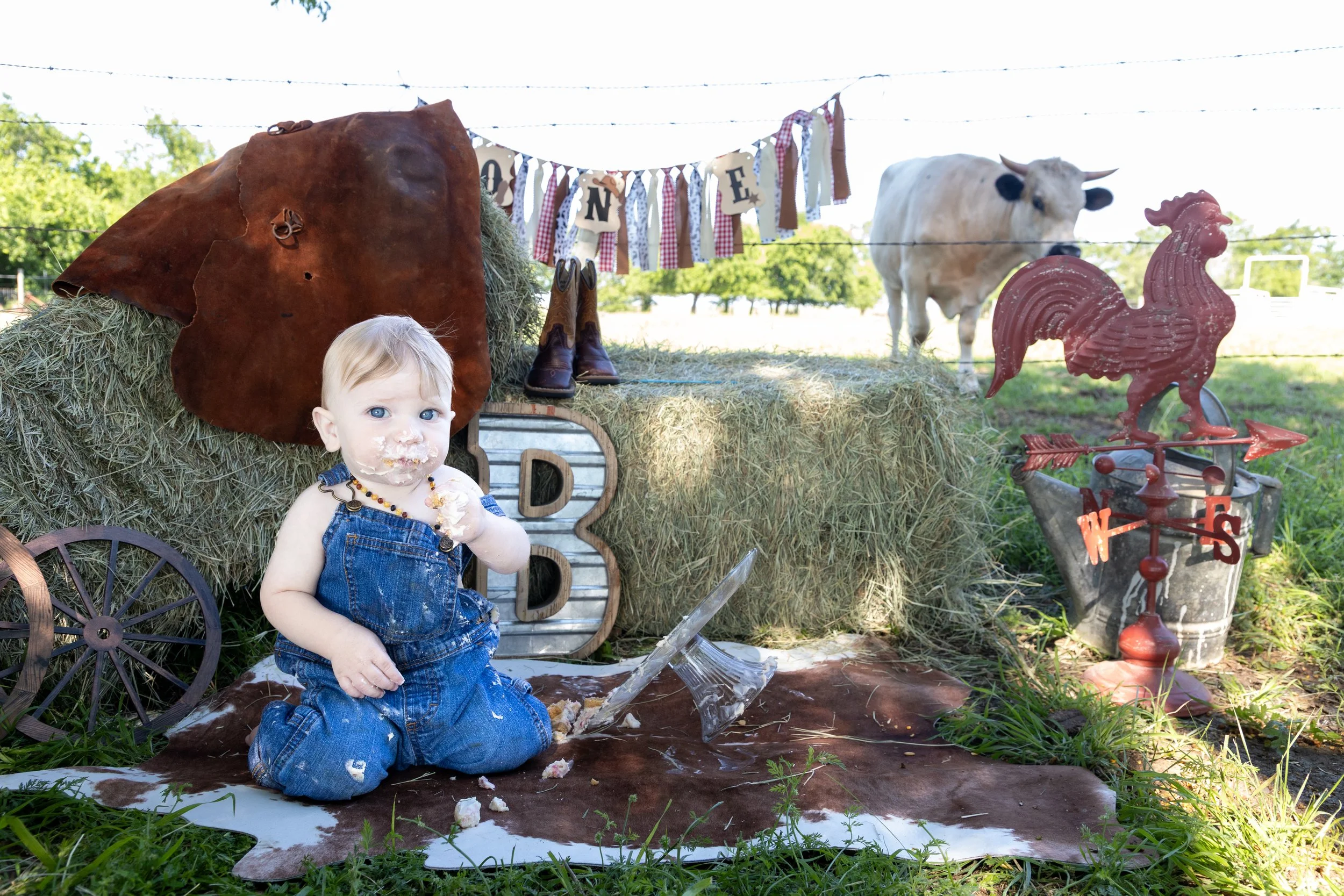 Child with blue eyes and blond hair, wearing denim overalls, sitting on a cowhide rug outdoors, covered in cake or pastry. Behind and around the child are hay bales, a vintage wagon wheel, and farm-themed decorations including a metal letter sign, a 