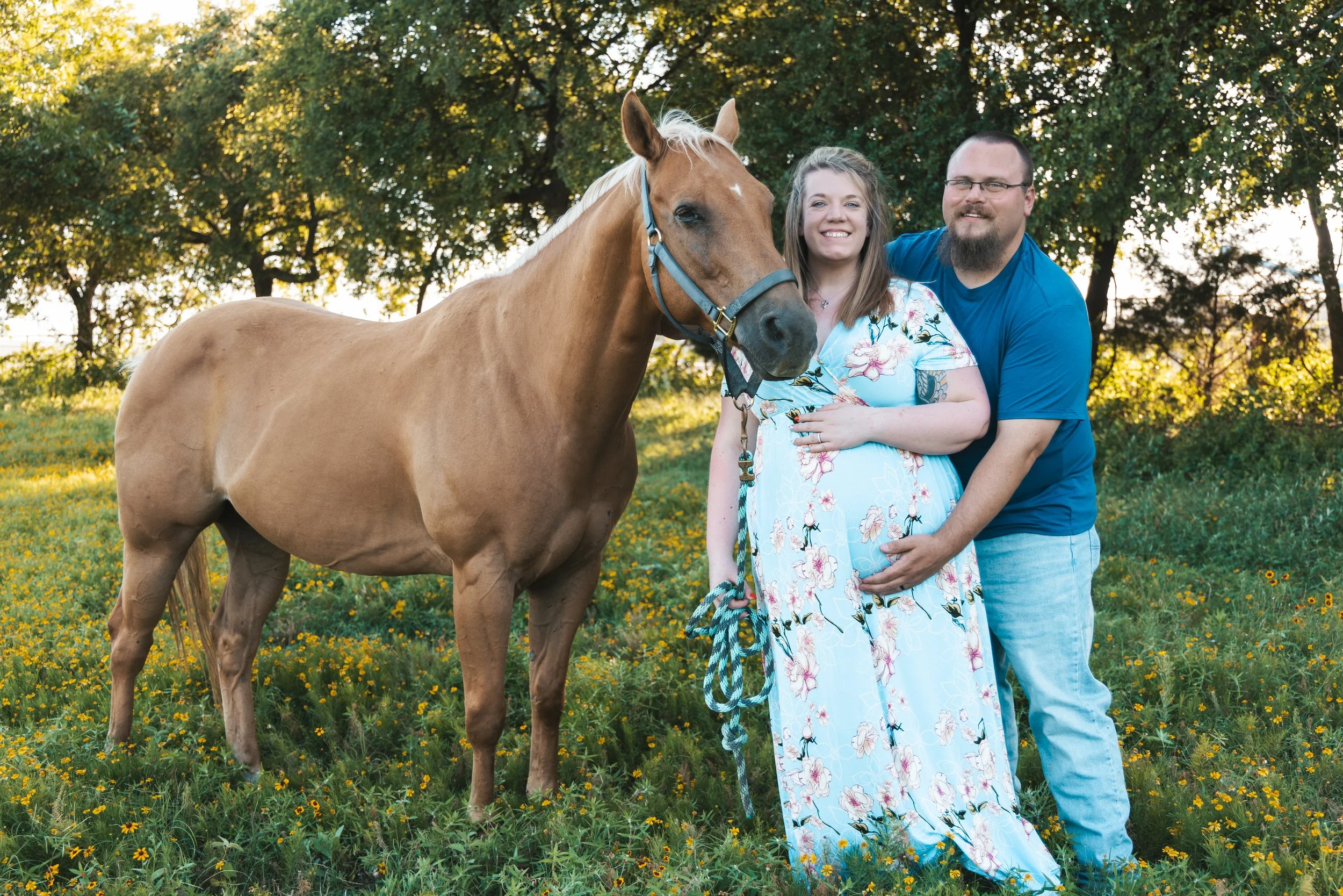 A smiling woman in a floral dress standing next to a man in a blue shirt, with a light brown horse, all outdoors in a grassy field with yellow flowers and trees in the background.
